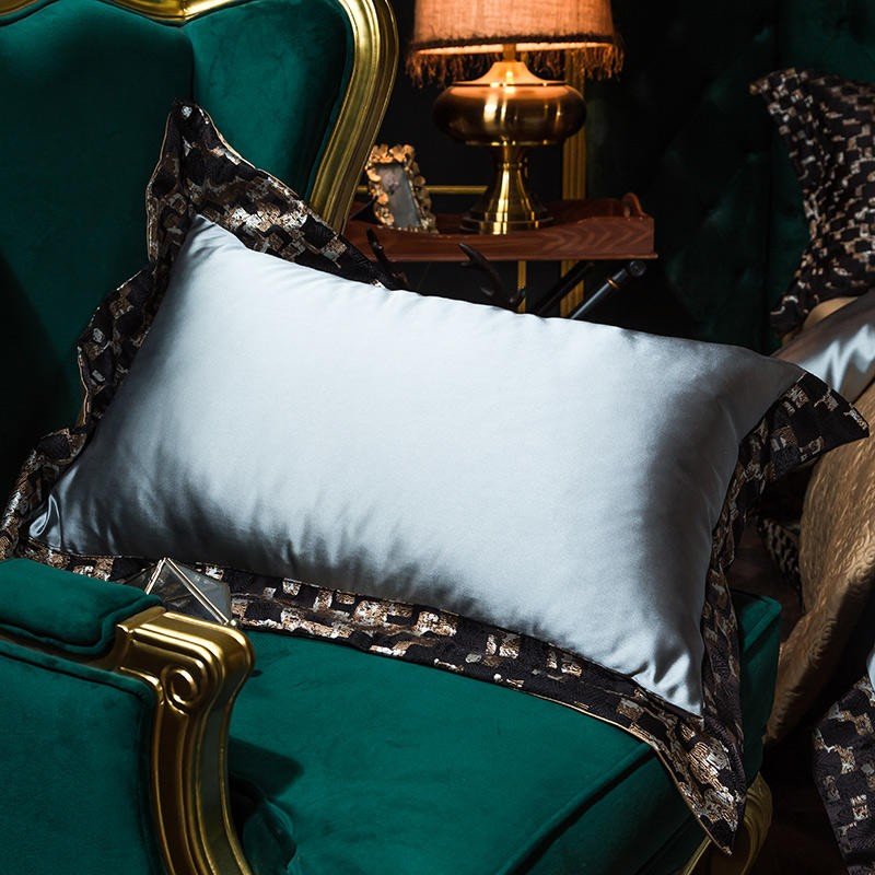 A close-up of a light blue satin pillowcase with a silky smooth finish, resting on a luxurious dark green velvet armchair. The pillow is accented with black and gold patterned trim, showcasing intricate geometric designs that enhance its opulent style. In the softly lit background, a brass lamp and wooden side table add warmth and elegance to the overall setting. The pillow's lustrous sheen highlights the premium Egyptian cotton fabric, reflecting light and adding sophistication to the scene.