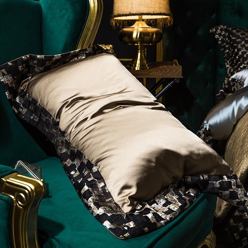A close-up of a satin pillowcase in a rich golden hue, placed on a dark green velvet armchair. The pillow is bordered with intricate black and gold patterned trim, adding a touch of elegance and sophistication. The smooth, luxurious texture of the fabric reflects light, emphasizing its premium quality. In the background, a warm-toned brass lamp and a hint of additional bedding contribute to the opulent ambiance of the scene.