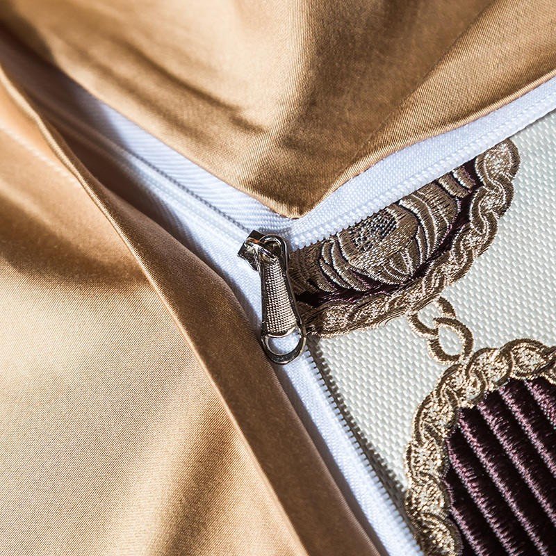A close-up of a golden satin bedding set featuring a white zipper closure, meticulously sewn into the fabric. The texture of the Egyptian cotton material is highlighted by its smooth, lustrous finish, reflecting light with a luxurious sheen. Adjacent to the zipper, an embroidered decorative pattern in gold and maroon threads adds intricate detail and a sophisticated touch to the bedding design. The craftsmanship showcases both durability and elegance, elevating the bedding’s opulent style.