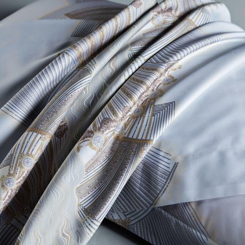 Close-up of an elegant white and beige bedding set featuring intricate gold and silver fan-like patterns with delicate swirling details. The luxurious fabric drapes softly, showcasing its smooth texture and ornate design.

