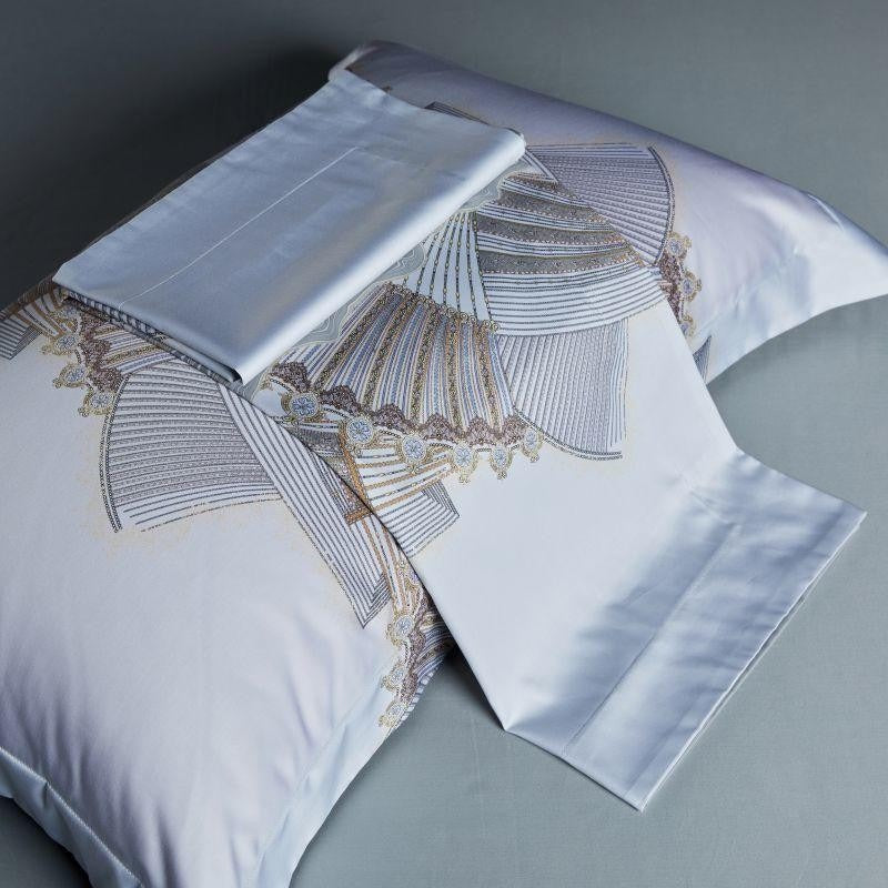 A pillowcase and matching folded flat sheet in soft white fabric with an intricate fan-like design in silver, gold, and beige hues, highlighted by elegant embroidered detailing.

