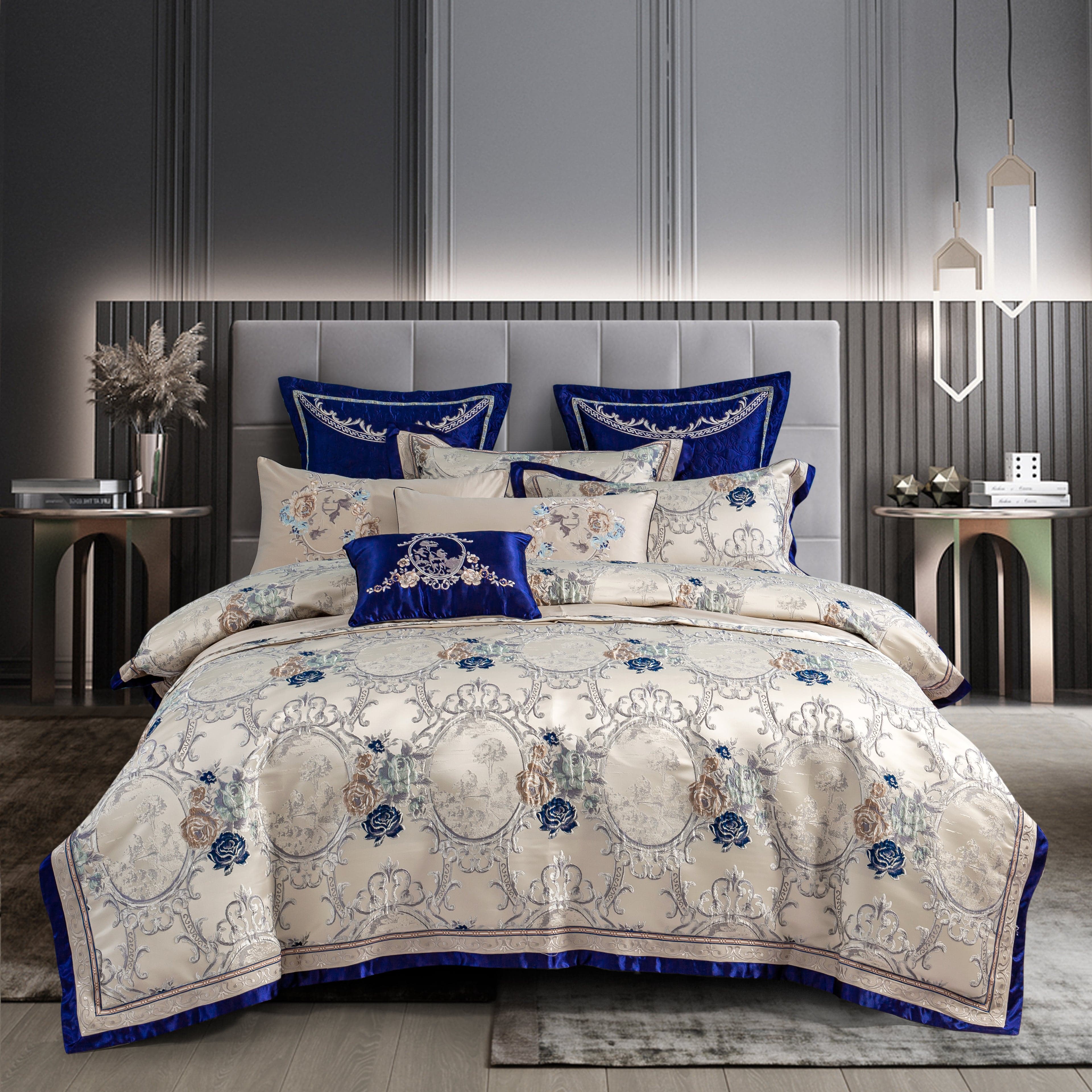 Luxurious bedding set with a cream-colored base featuring intricate floral and ornate patterns in blue and silver tones, accented by royal blue velvet pillows and trim. The elegant design complements the modern, neutral-toned bedroom setting.