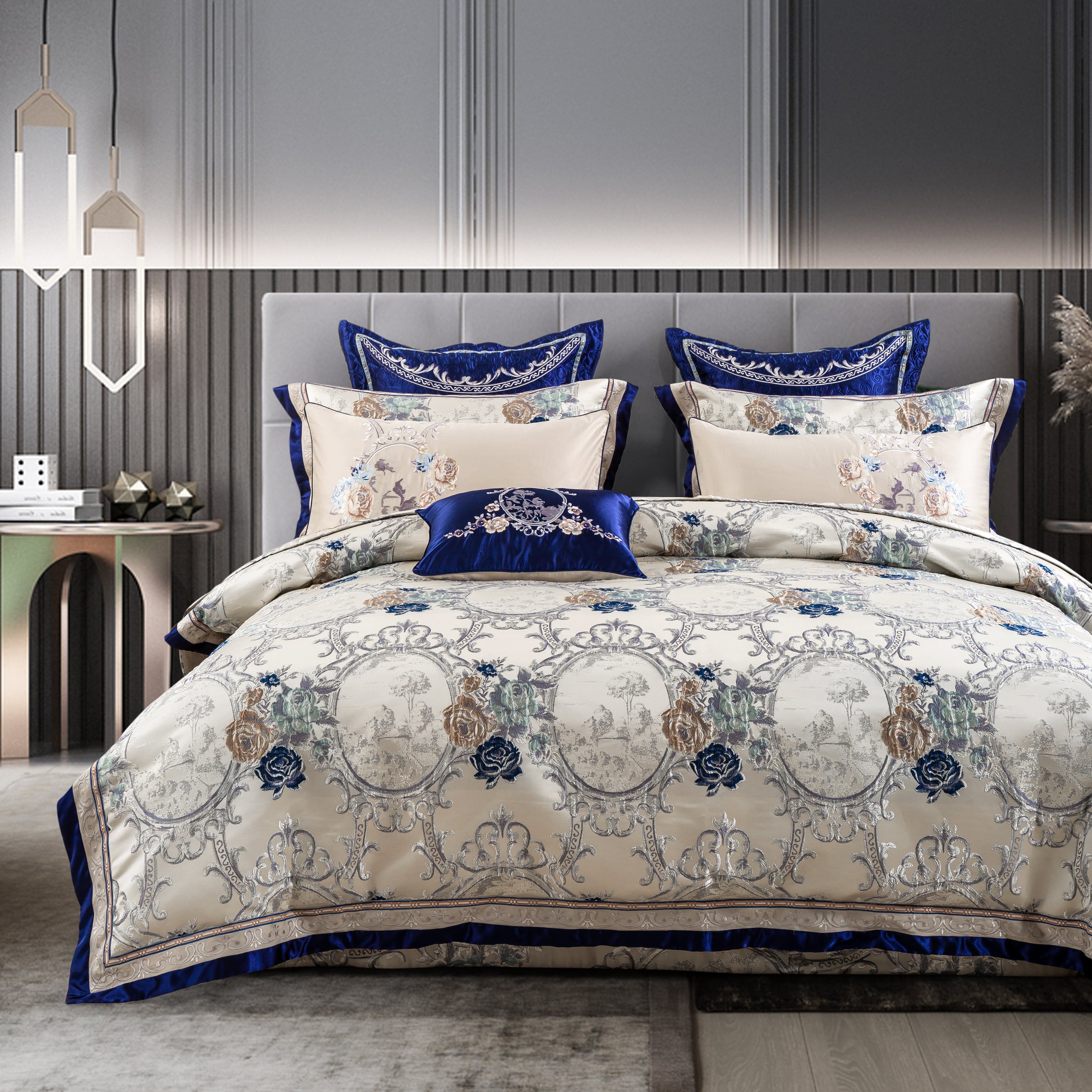 Bedding set with a luxurious cream base featuring ornate floral and decorative motifs in blue and silver tones, enhanced by royal blue velvet accents and a matching decorative pillow. The set is displayed in a modern bedroom with a sleek gray upholstered headboard and elegant side tables.