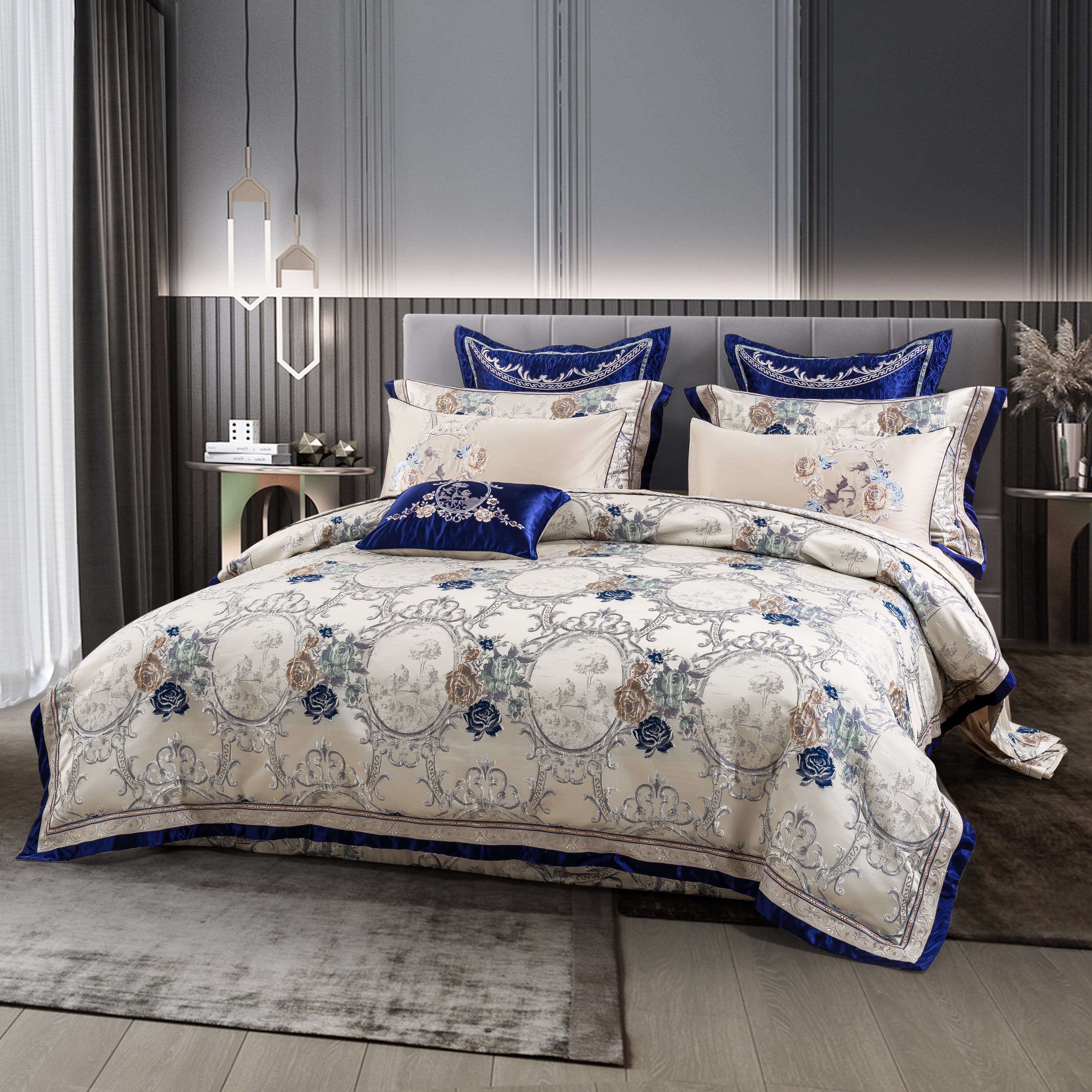 Elegant bedding set with a cream-colored base adorned with intricate floral and decorative patterns in shades of blue and silver, complemented by royal blue velvet accents and pillow trims. The sophisticated design harmonizes with the contemporary bedroom featuring soft lighting and a neutral palette.