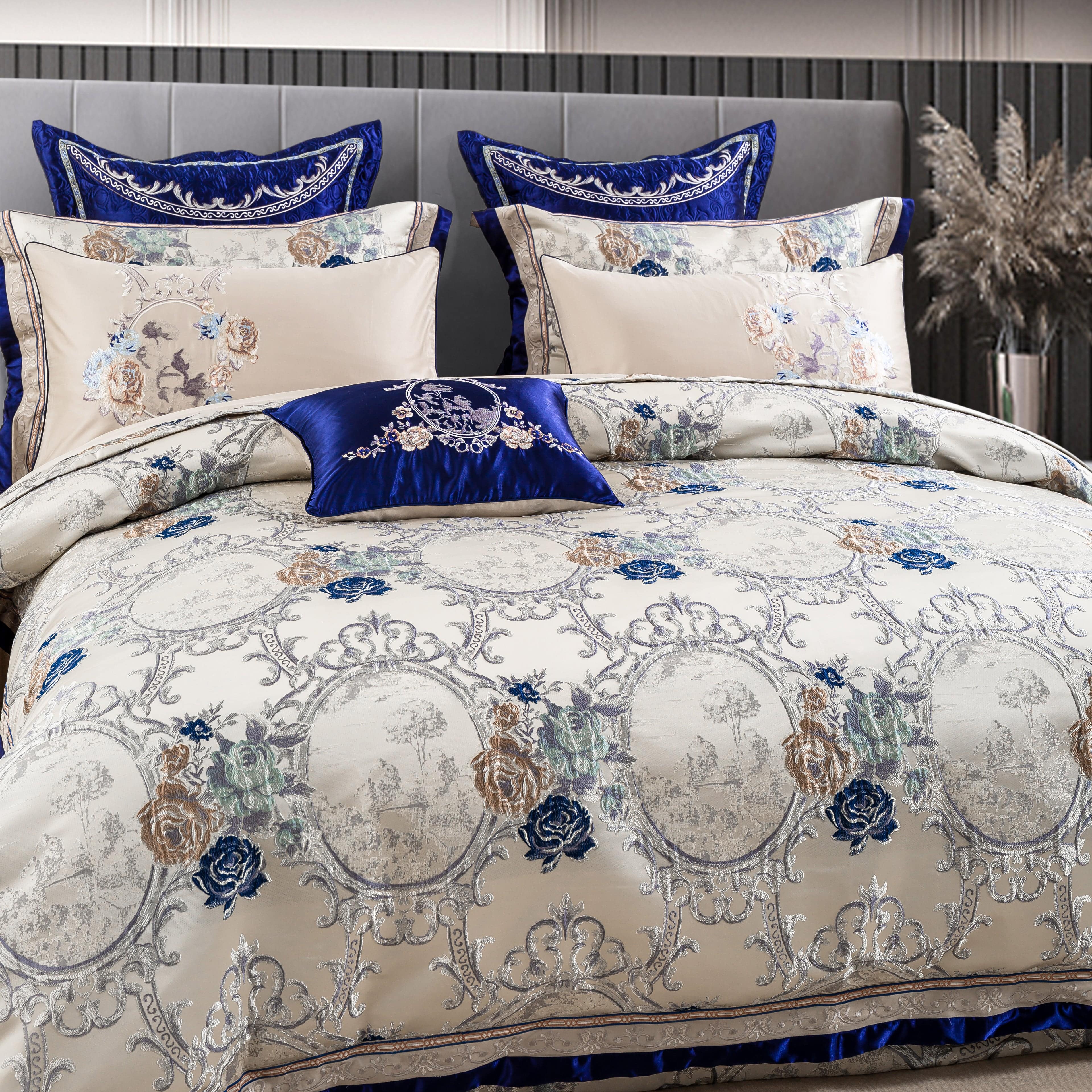 An elegant bedding ensemble featuring a luxurious cream duvet cover adorned with intricate silver and blue floral medallion patterns. Matching pillows, bordered with royal blue satin accents and detailed embroidery, complement the design. A decorative royal blue accent pillow with embroidered white floral motifs completes the sophisticated look.