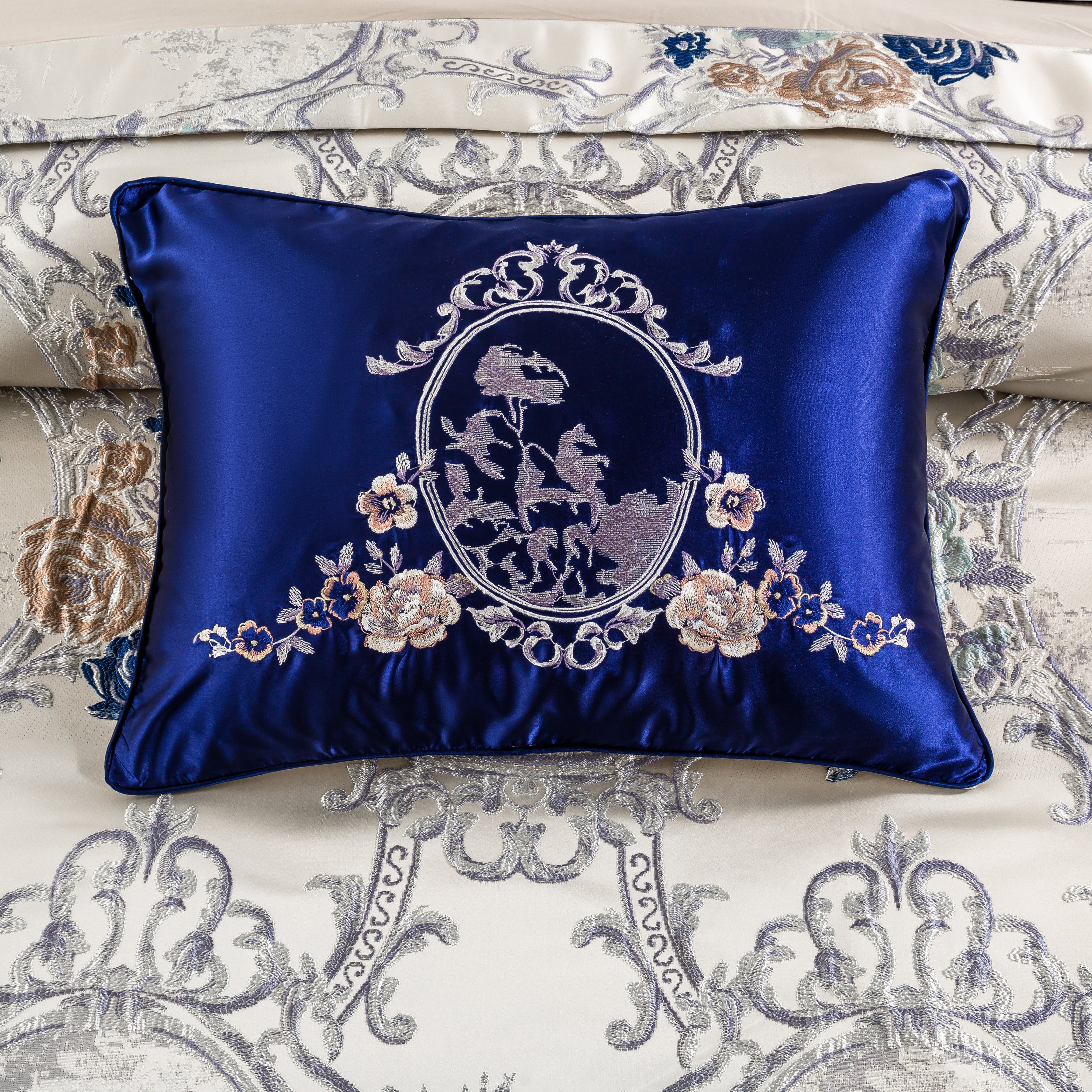 A close-up Intricate baroque-inspired pattern featuring a central oval frame with a subtle pastoral scene surrounded by ornate silver filigree. Vibrant floral accents of blue, brown, and green roses add contrast and elegance to the cream backview of a decorative royal blue satin pillow featuring intricate silver embroidery of a romantic equestrian scene framed by ornate floral and scrollwork designs. Below the frame, delicate embroidered floral arrangements in soft gold and blush hues add a luxurious touch.