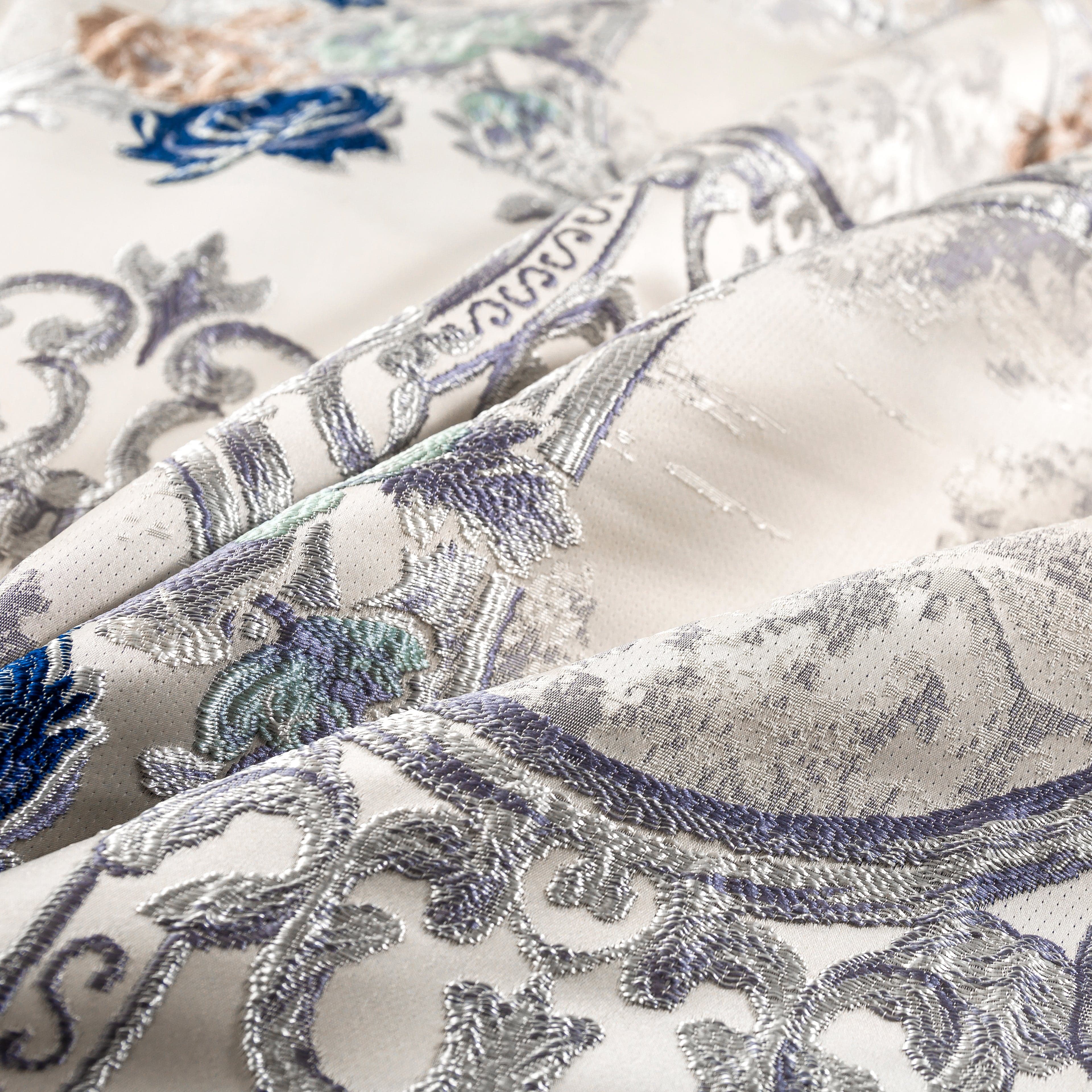 Close-up of an intricate jacquard fabric featuring a detailed floral and ornamental pattern in silver, blue, and pastel tones on a cream background.