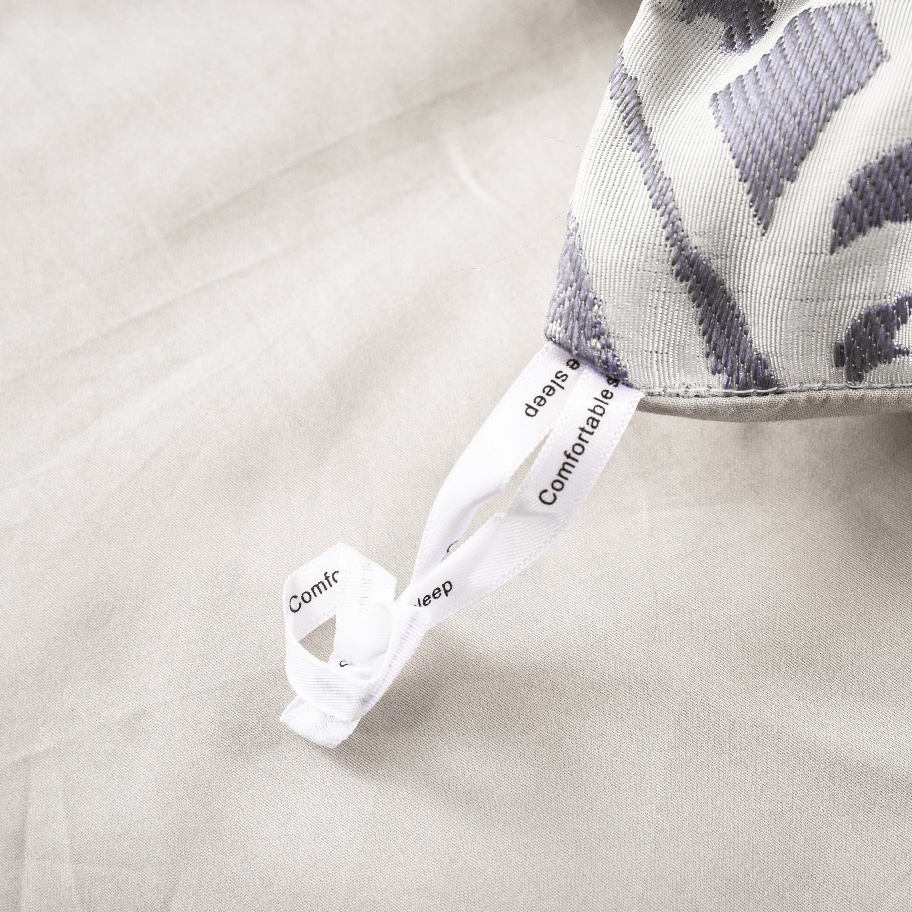 Close-up of a fabric corner with a white ribbon loop labeled "Comfortable Sleep," designed for securing bedding in place. The textured fabric features a subtle gray pattern on a neutral base.