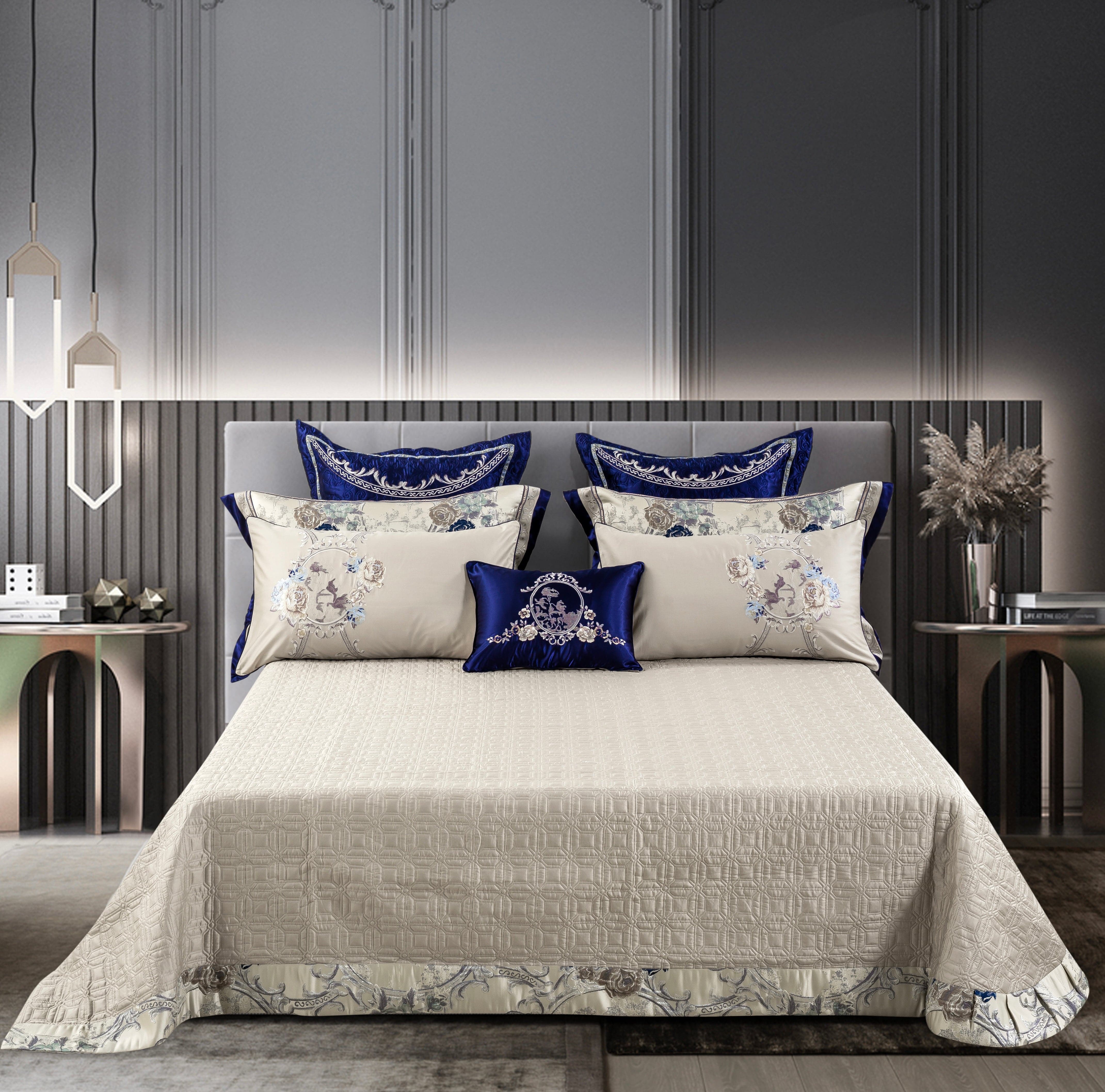 A lavishly designed bedding set featuring a smooth cream-colored bedspread with intricate floral and ornamental patterns along the hem. The bed is dressed with matching pillow shams and decorative cushions in ivory and royal blue, highlighted by elegant embroidery. The minimalist yet luxurious setting includes contemporary side tables and decor, creating a modern and opulent bedroom ambiance.