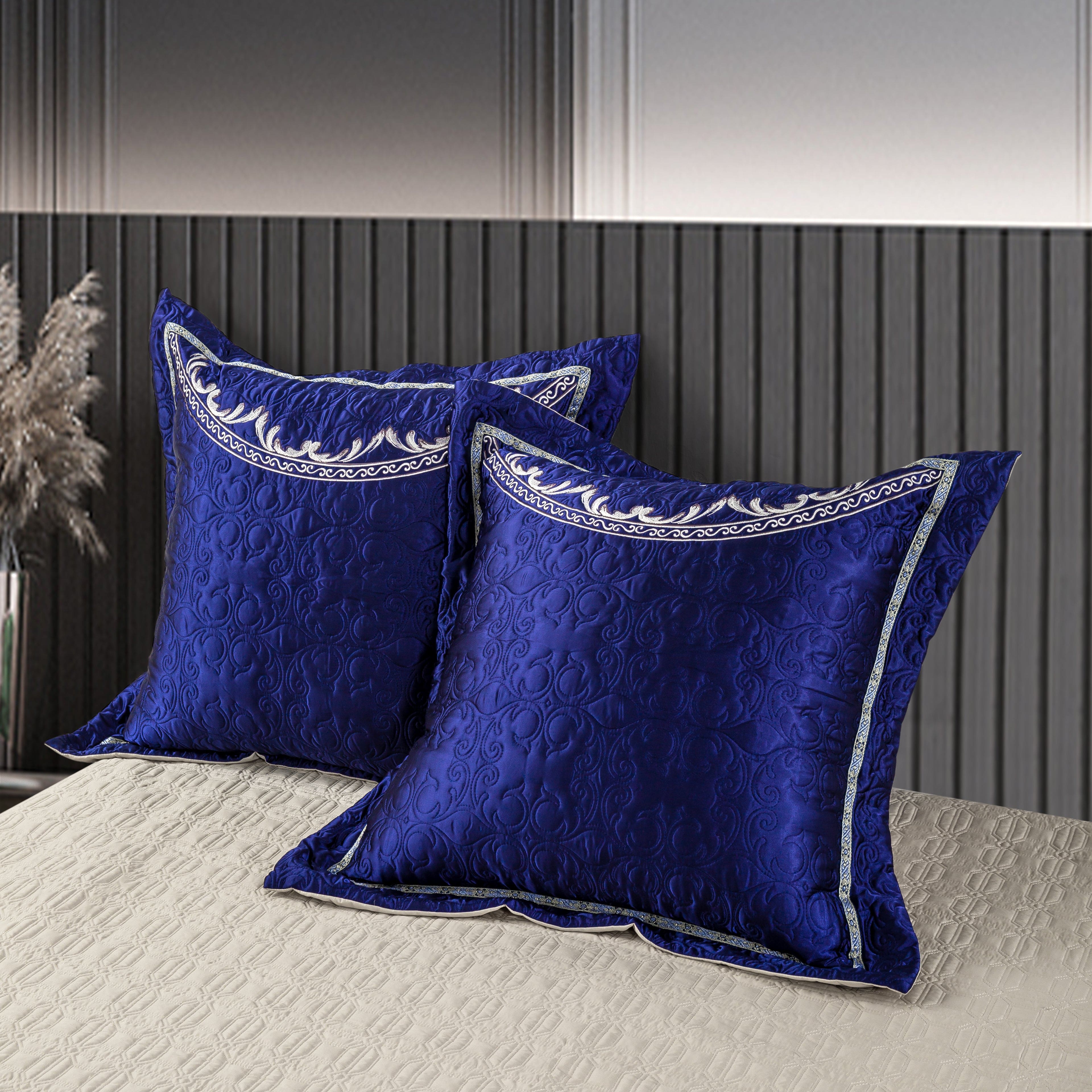 Two square blue pillowcases with a luxurious quilted texture, bordered by intricate silver embroidery in a swirling pattern, resting on a textured cream bedspread against a modern paneled wall background.