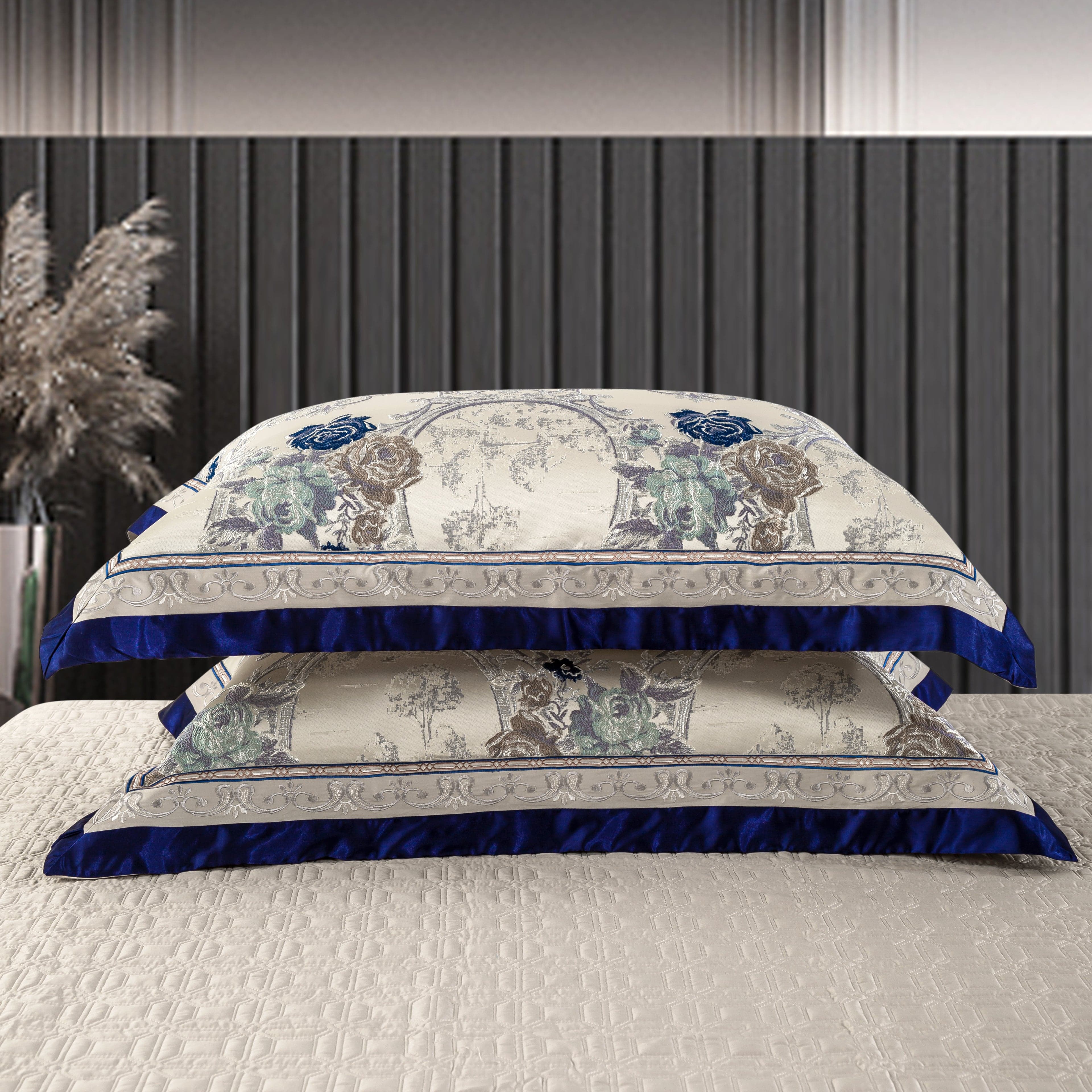 Stacked decorative pillows featuring a cream base with intricate floral patterns in blue and silver tones, bordered by vibrant royal blue satin accents. The pillows are placed on a textured cream bedspread in a sophisticated modern bedroom setting.