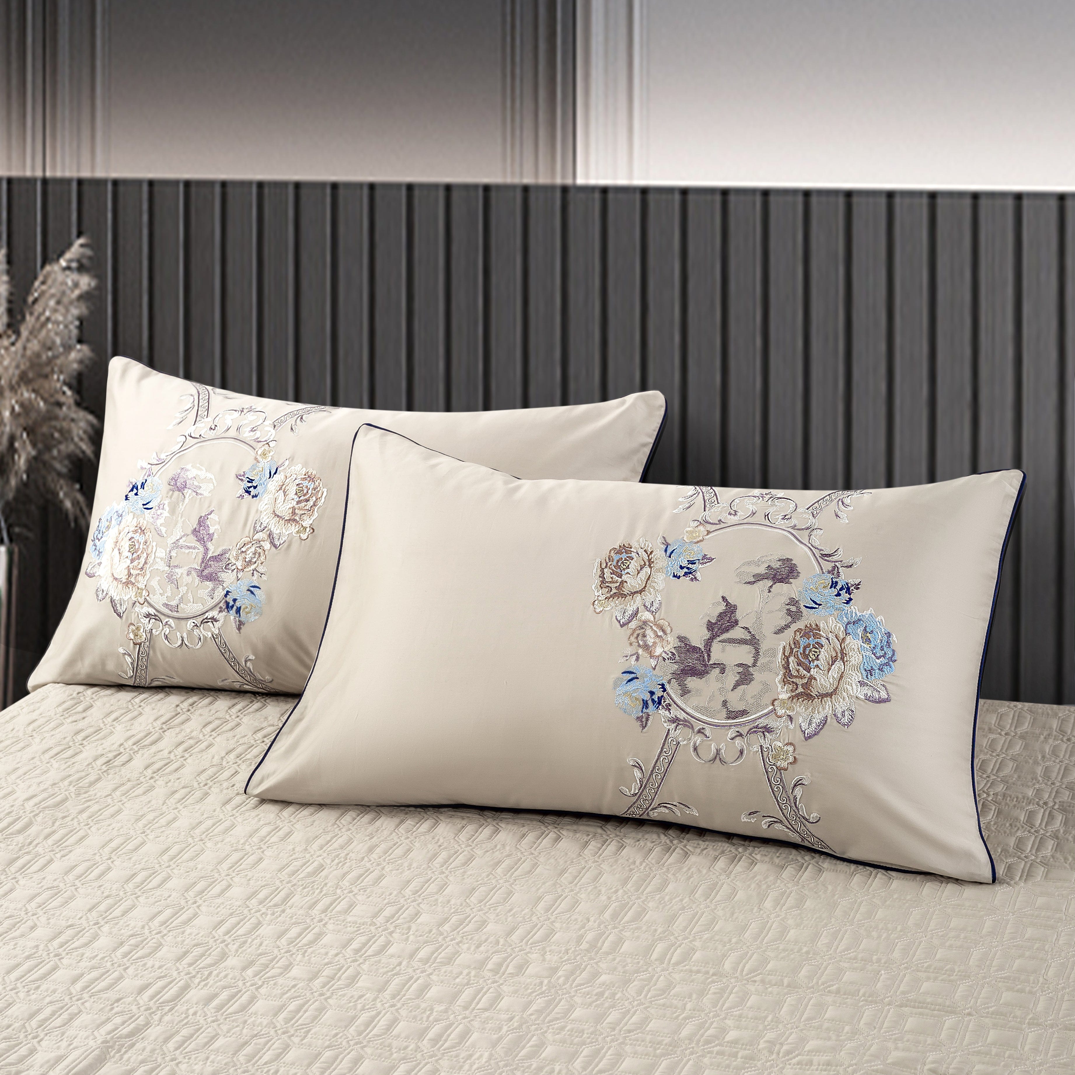 Two elegant beige pillowcases adorned with intricate floral embroidery in muted gold and blue tones, showcasing a central circular motif featuring delicate designs. The pillows rest on a textured cream quilted bedspread, complementing the classic and luxurious bedroom decor.