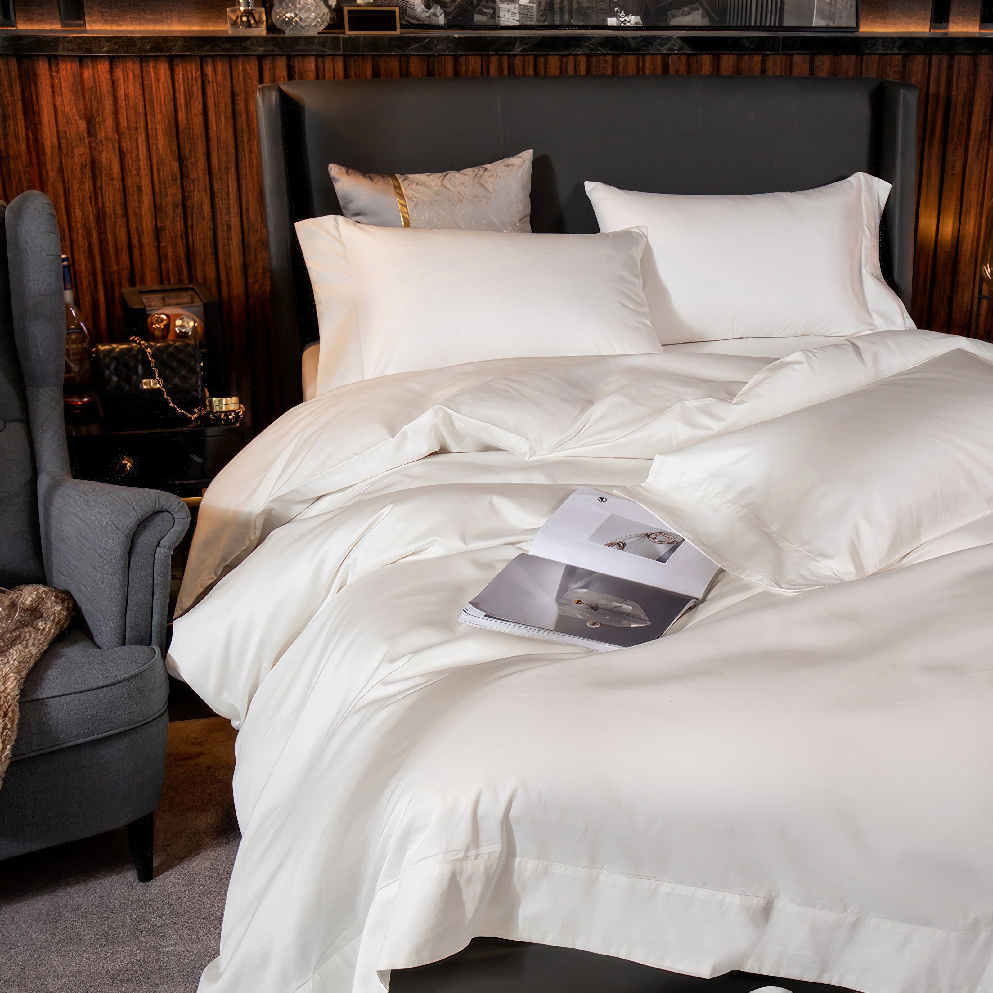 A close-up view of a pristine white bedding set with a smooth, silky finish, featuring neatly arranged pillows and a draped duvet. A magazine is casually placed on the bed, adding a touch of lifestyle appeal. The scene is complemented by a chic urban backdrop with warm wood paneling, a grey armchair, and sophisticated bedside accessories, evoking a luxurious and inviting atmosphere.

