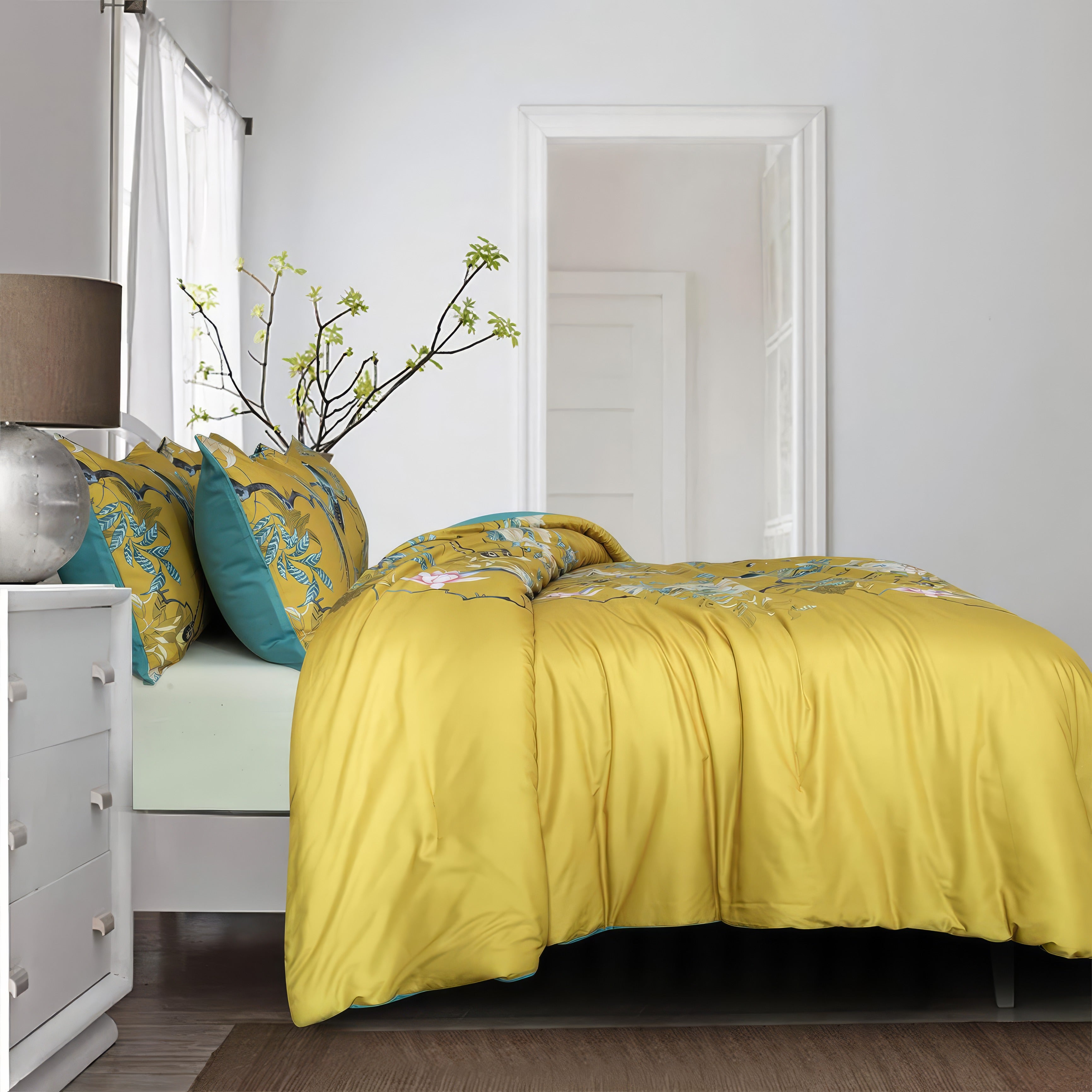 A modern bedroom featuring a mustard yellow duvet cover with intricate floral and bird patterns, paired with matching pillowcases backed by solid teal accents. The bed is framed by a clean, minimalist white dresser topped with a metallic lamp and a vase holding delicate branches with green blooms. A large mirror reflects light from a nearby window, enhancing the airy ambiance, while a soft beige rug anchors the space. The white walls and natural decor elements create a serene, inviting atmosphere.