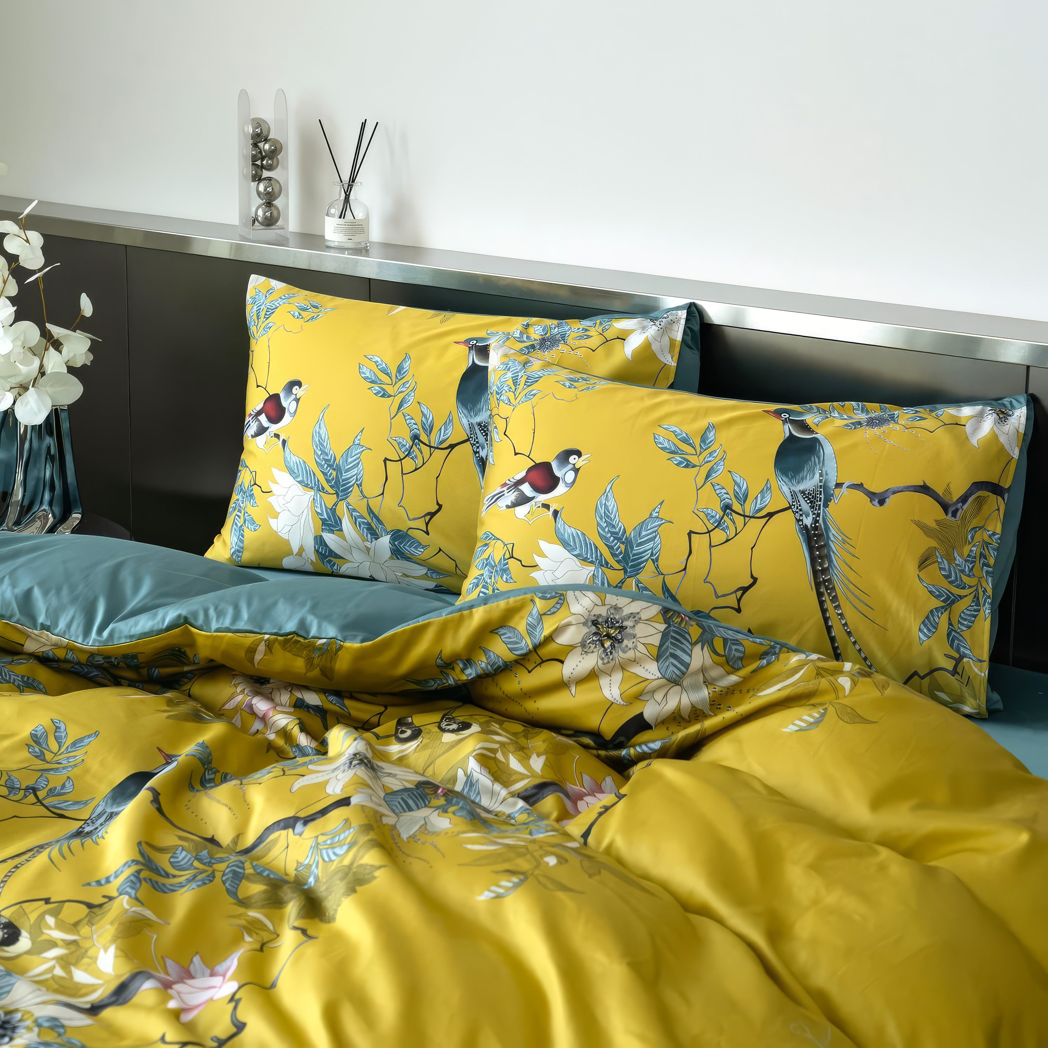 A stylish bedroom setting showcasing a mustard yellow bedding set with vibrant floral and bird motifs, paired with teal accents on the reverse side of the comforter. The pillowcases feature intricate artwork of birds perched on branches amidst blooming flowers and lush leaves. A sleek metallic headboard adds a contemporary touch, while decorative elements like a vase with white flowers and a diffuser on the shelf complete the serene, modern aesthetic.