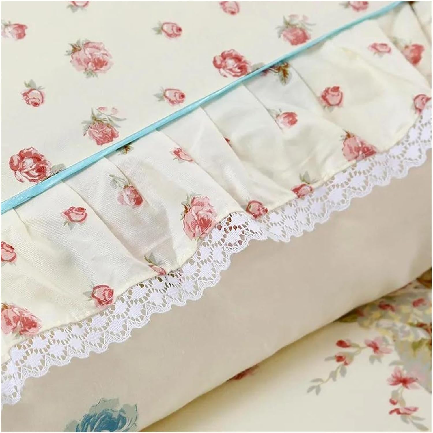 A close-up of cream-colored bedding with a delicate floral pattern of small red and pink roses scattered evenly. A light blue trim runs along the edge, separating the main fabric from a ruffled section adorned with matching floral designs. Beneath the ruffles, intricate white lace detailing adds a soft, elegant finish. The fabric is smooth and slightly glossy, highlighting its refined quality.

