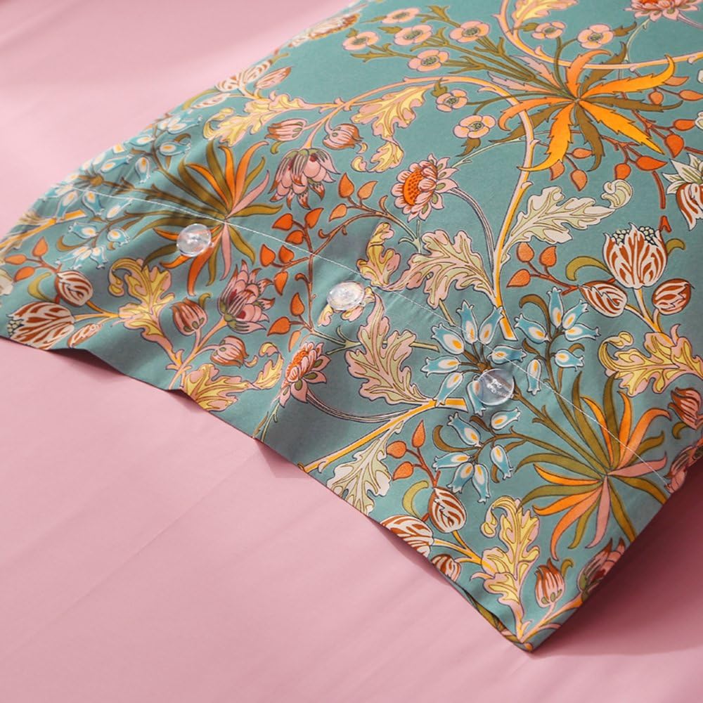 A close-up of a decorative pillowcase featuring a detailed floral pattern with vibrant orange, pink, and blue flowers and leaves on a muted teal background. Transparent buttons line the edge, adding a functional yet elegant detail. The pillowcase rests on a soft pink bedsheet, creating a visually appealing contrast between the bold design and solid background. The fabric appears smooth and luxurious, enhancing the artistic and vintage aesthetic.

