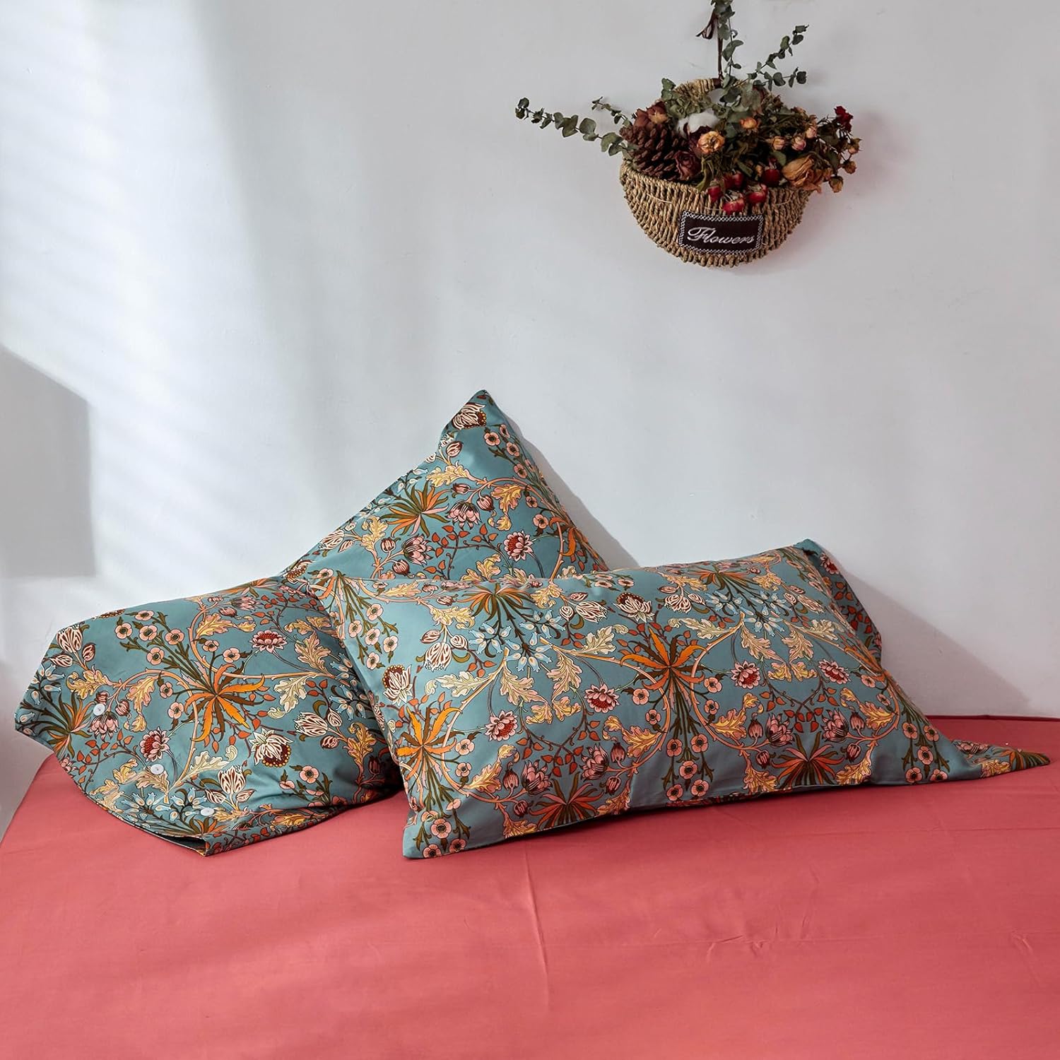 Two decorative pillowcases with an intricate floral design featuring orange, red, and beige patterns on a muted teal background. The symmetrical botanical motifs include blooming flowers, leaves, and branches, adding a vintage yet vibrant aesthetic. The pillows rest on a smooth coral-red bedsheet, which contrasts beautifully with the detailed design. A woven basket filled with dried flowers and greenery hangs on the white wall above, enhancing the cozy and artistic ambiance.

