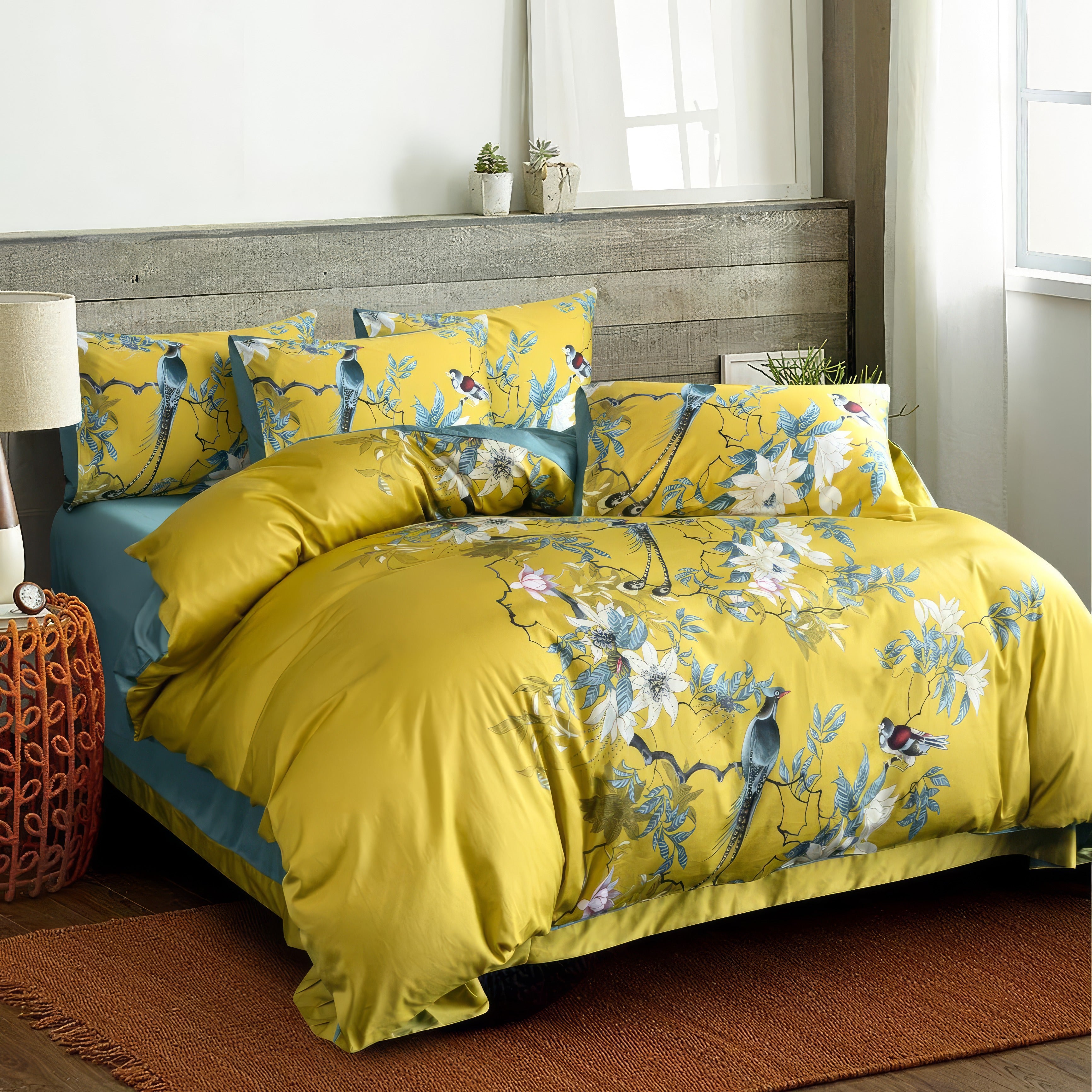 A bright and lively bedding set showcasing a mustard yellow duvet cover and matching pillowcases adorned with intricate bird and floral designs in blue, white, and red. The bed is placed in a rustic bedroom featuring a wooden plank headboard, a woven bedside table with a clock, and a large window allowing natural light. A textured orange rug adds warmth to the decor, complemented by potted plants on the headboard shelf.