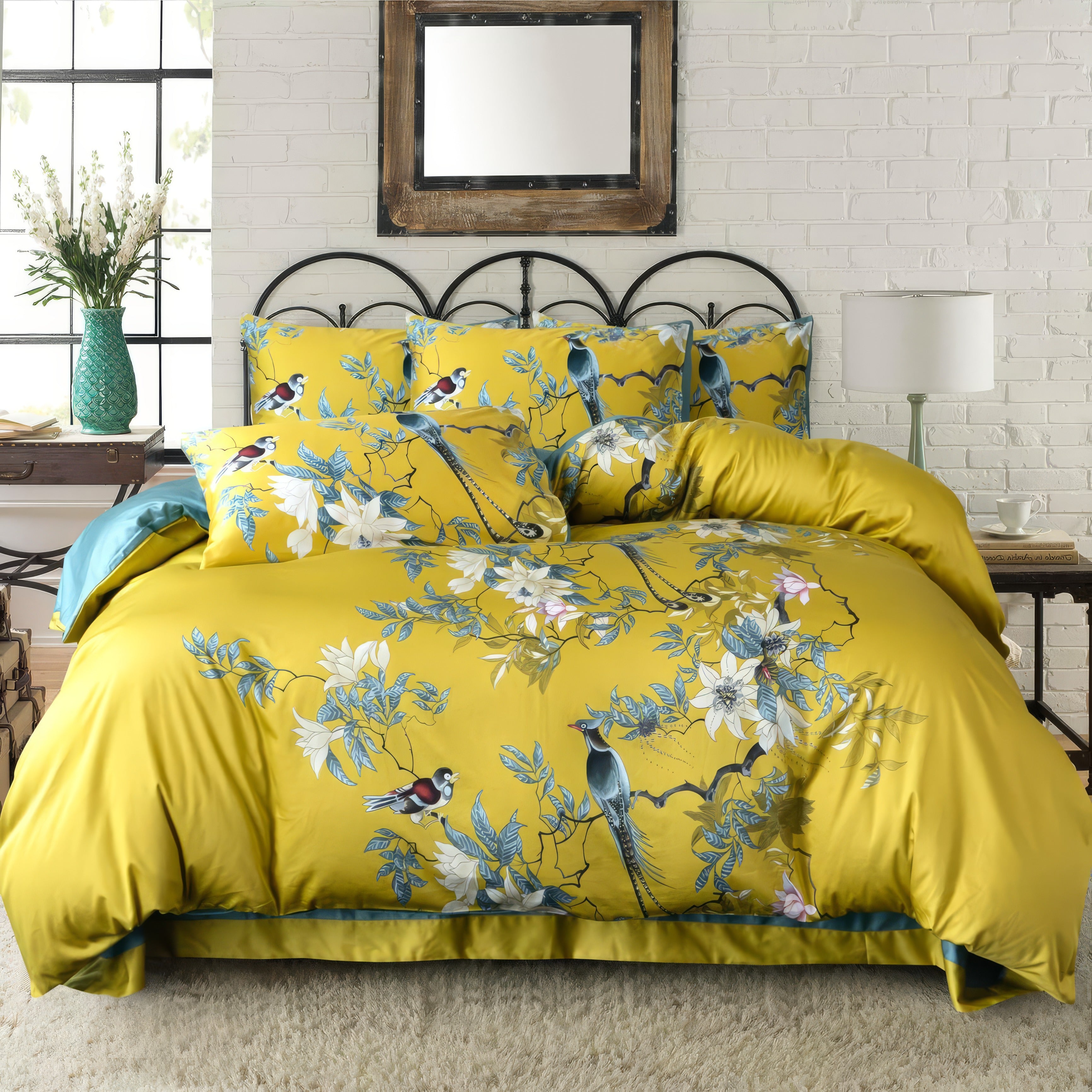 A vibrant bedding set featuring a gold duvet cover and pillowcases adorned with intricate bird and floral prints in blue, white, and red. The bed is styled in a cozy farmhouse bedroom with a wrought-iron headboard, white brick wall, and rustic wooden mirror. A green vase with fresh flowers and a white bedside lamp complement the setting.






