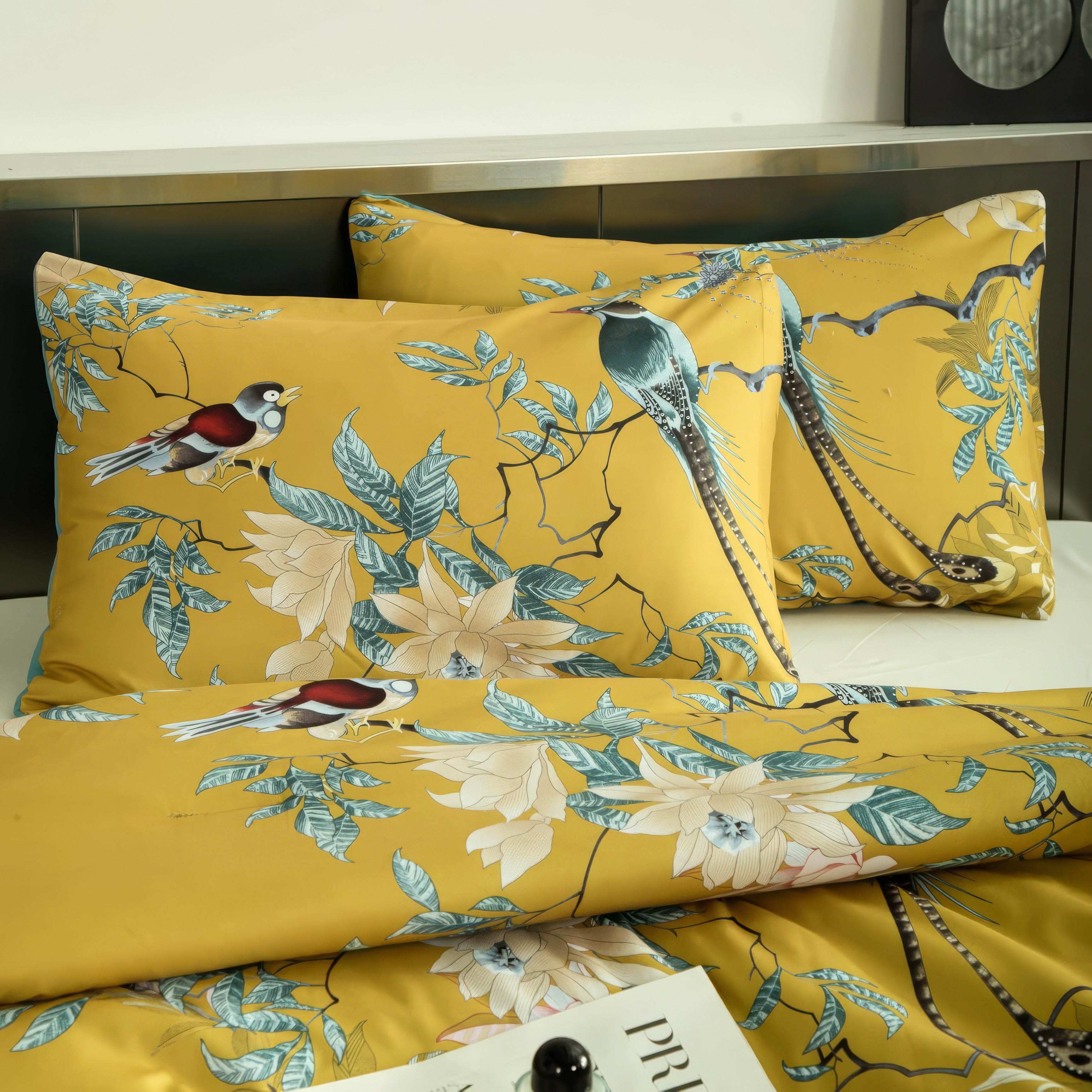 Close-up of a mustard yellow bedding set featuring detailed illustrations of birds perched on tree branches with blooming flowers and vibrant green leaves. The pillowcases perfectly align with the intricate floral and bird motifs, creating a cohesive and elegant design. The smooth fabric showcases a luxurious sheen, complemented by a minimalist metallic background and a magazine placed on the bed for a modern, refined aesthetic.