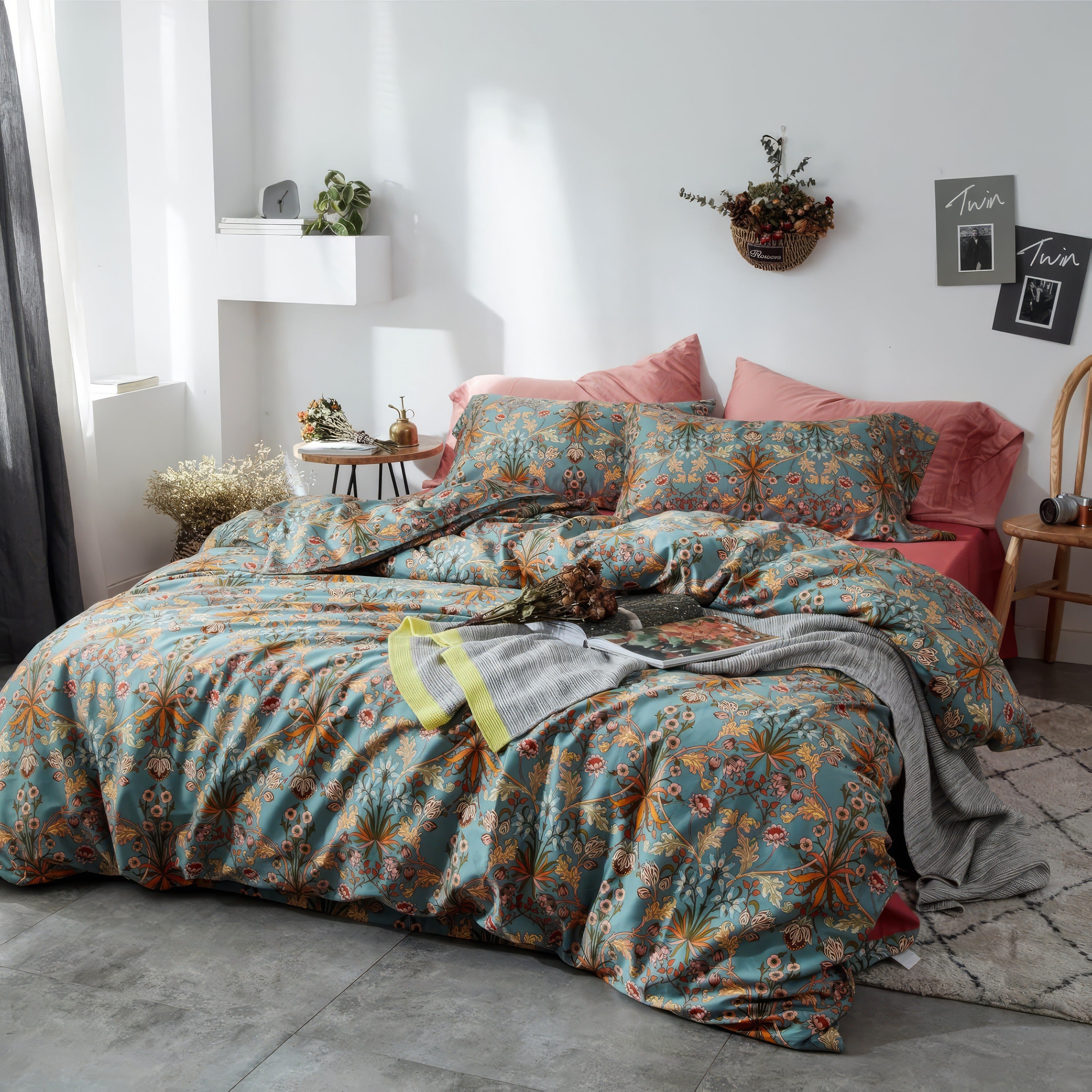 A cozy bedroom featuring a botanical-themed bedding set in soft teal with intricate orange, yellow, and white floral patterns. The duvet and matching pillowcases are paired with two solid blush pink pillows against a white wall. The decor includes a wooden chair, a hanging wall basket filled with dried flowers, and framed black-and-white prints. Natural light fills the space, enhancing the warm and inviting atmosphere.

