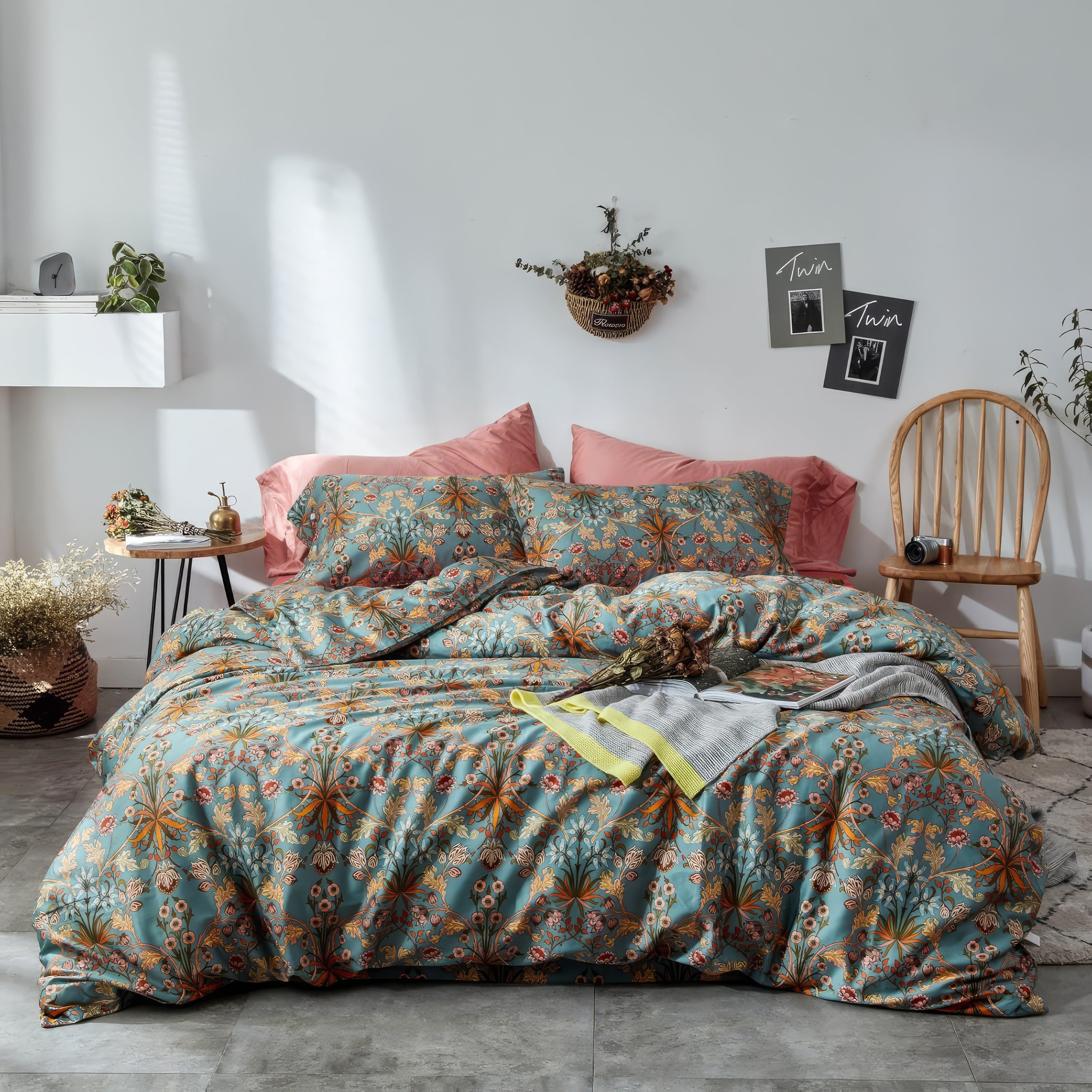 Botanical-themed bedding set in soft teal adorned with intricate orange, yellow, and white floral patterns. The duvet and matching pillowcases are paired with two solid blush pink pillows against a white wall. A wooden chair, hanging wall basket with dried flowers, and framed black-and-white prints decorate the cozy bedroom, with a natural light cast over the space.

