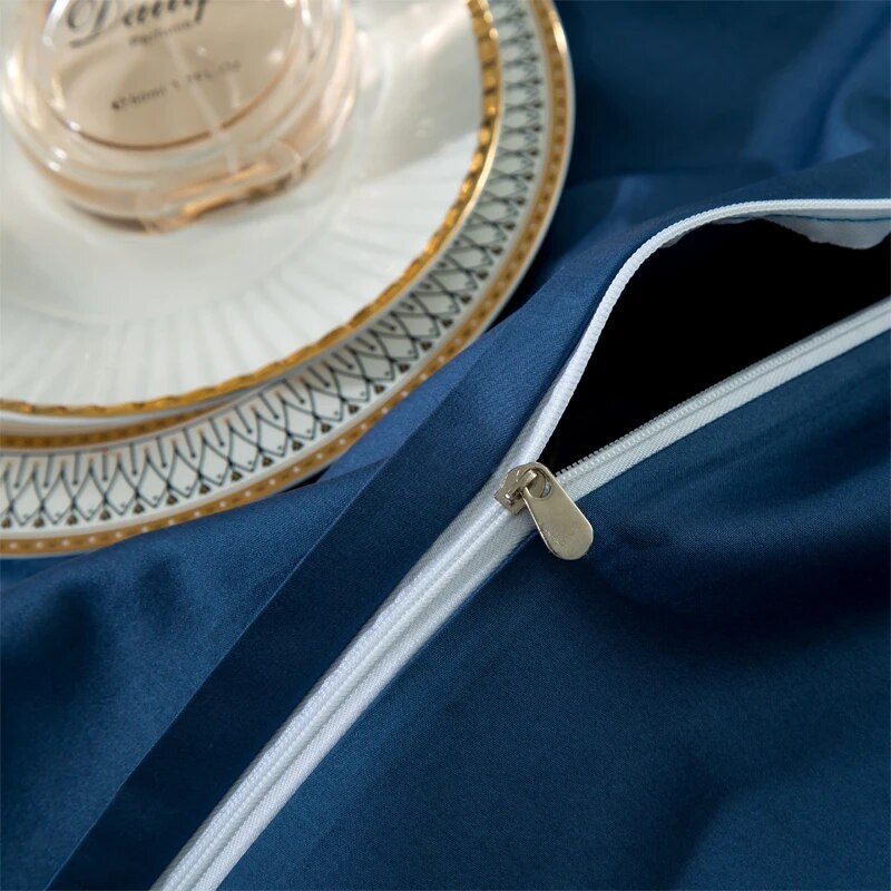 Close-up of a blue bedding fabric featuring a white zipper with a silver pull tab. A white ceramic plate with intricate gold detailing rests on the bedding, along with a clear glass perfume bottle, adding a touch of elegance and luxury. The smooth fabric and precise stitching highlight the quality of the bedding.

