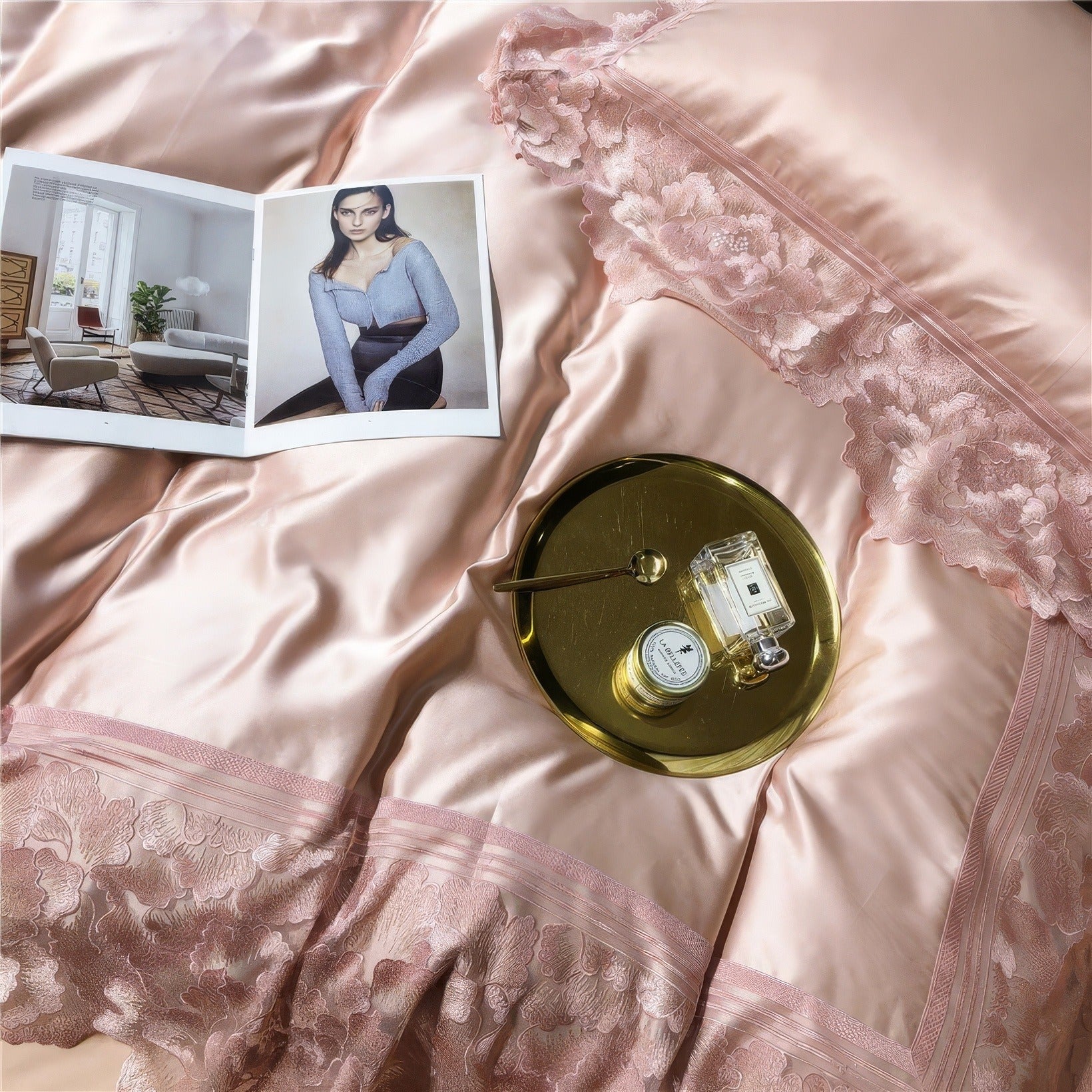 Pink satin bedding with floral lace detailing, an open magazine displaying a model in a blue outfit, and a gold tray holding a perfume bottle, cream jar, and a spoon.

