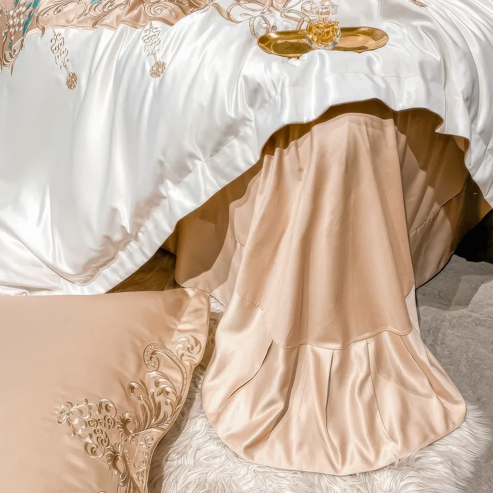 A golden beige bed skirt with a tiered, pleated design cascading to the floor, complementing a luxurious white duvet cover adorned with intricate golden embroidery. The setup exudes elegance, with the soft shimmer of the fabric enhancing the sophisticated bedding ensemble.

