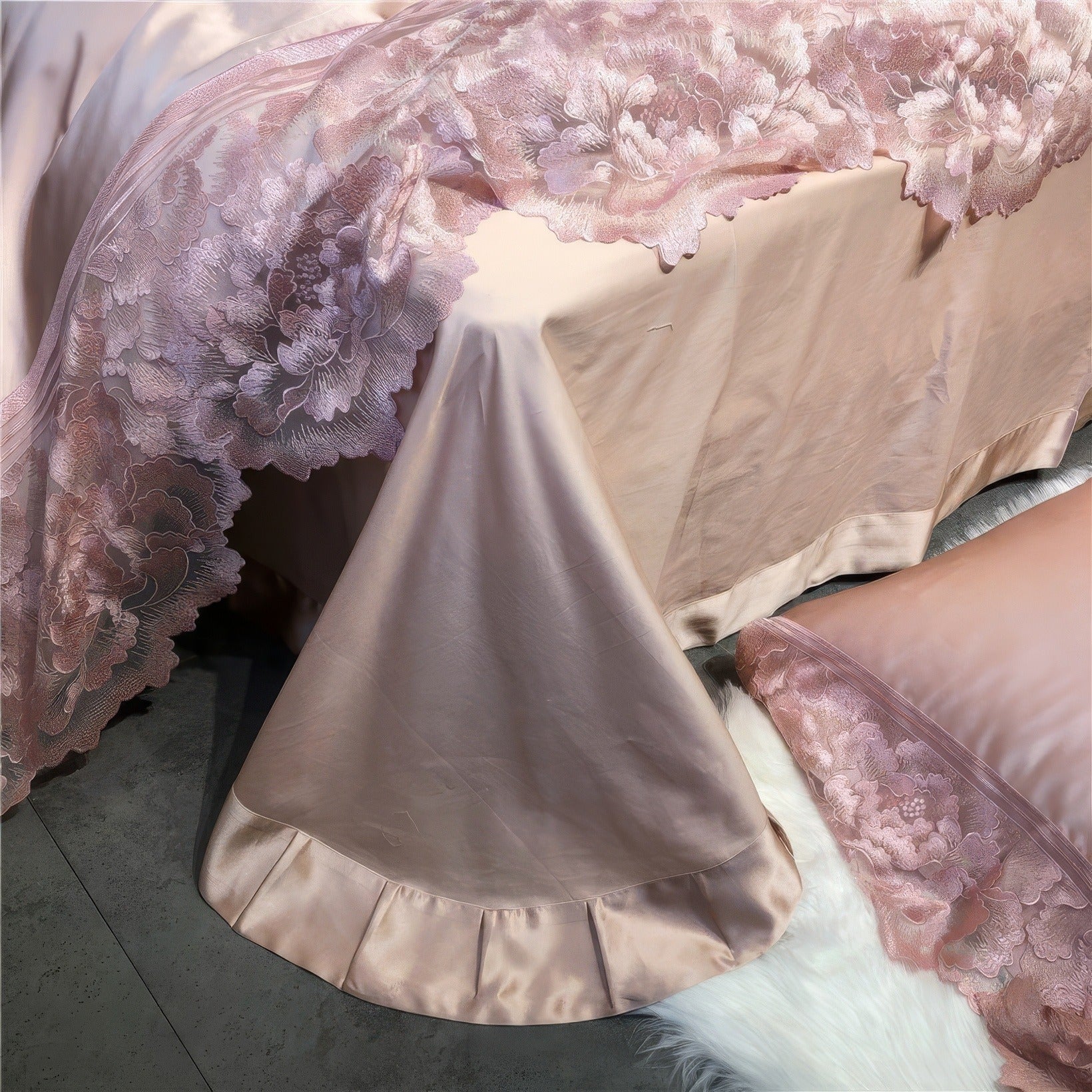 Close-up of pink satin bedding featuring intricate floral lace trim along the edges and a pleated skirt detail draped over the side of the bed. A matching pink satin pillowcase with lace trim rests on a soft white rug.

