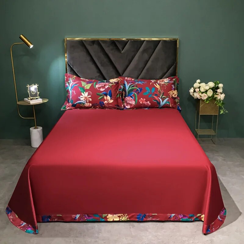 Luxurious crimson Egyptian cotton bedding set featuring a smooth solid red sheet accented with vibrant floral prints at the edges. Matching pillowcases with intricate multicolored botanical designs complete the elegant setup, complemented by a plush velvet headboard and soft lighting in a contemporary bedroom setting.

