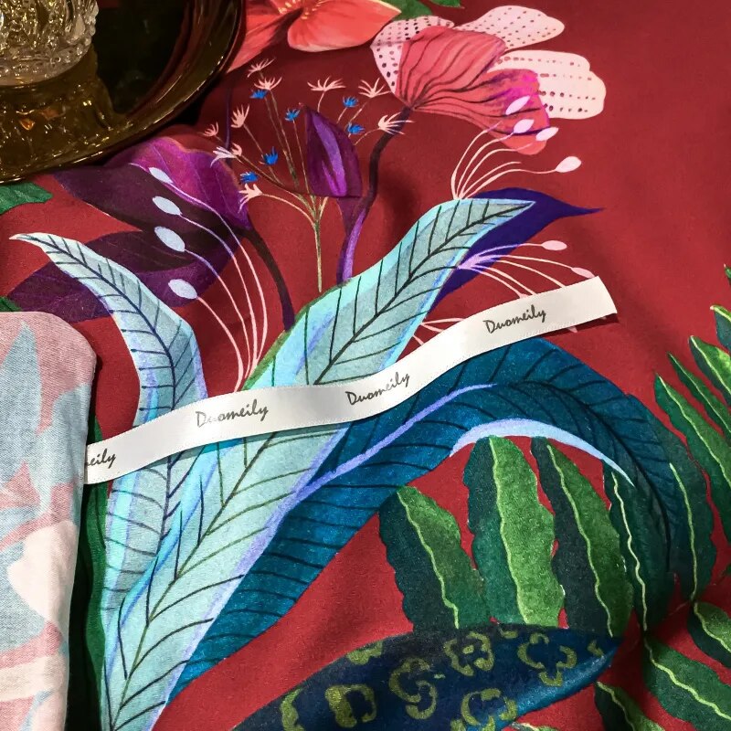 Close-up of a crimson Egyptian cotton fabric with vivid floral and botanical patterns in shades of blue, green, and purple. A white ribbon labeled "Duomily" rests atop the fabric, next to a polished gold tray with a textured crystal jar, adding an element of sophistication.

