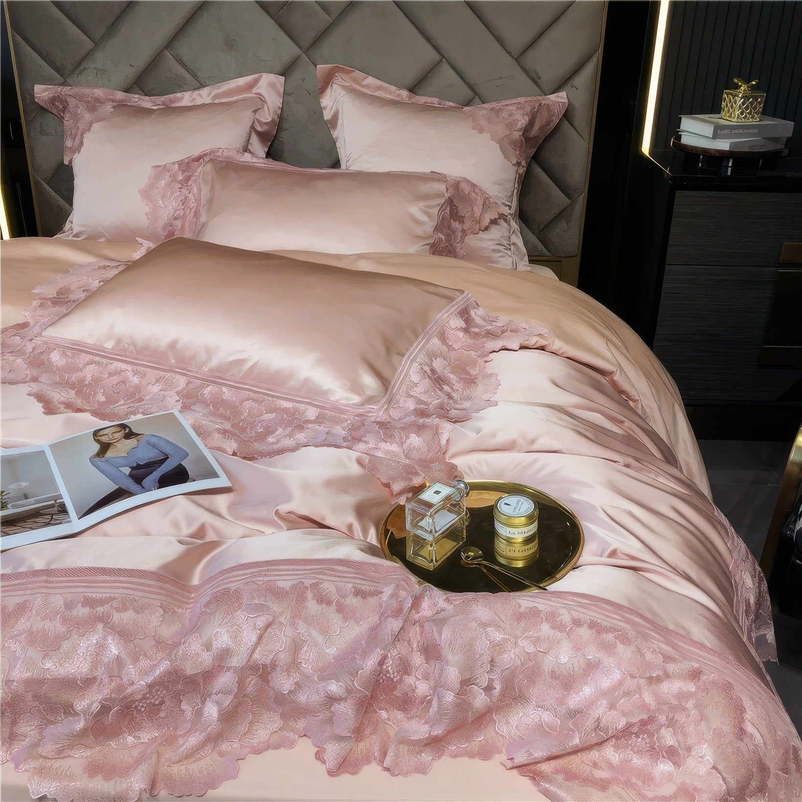 Pink satin bedding adorned with intricate floral lace details, accompanied by a gold tray holding a perfume bottle, cream jar, and a small spoon.

