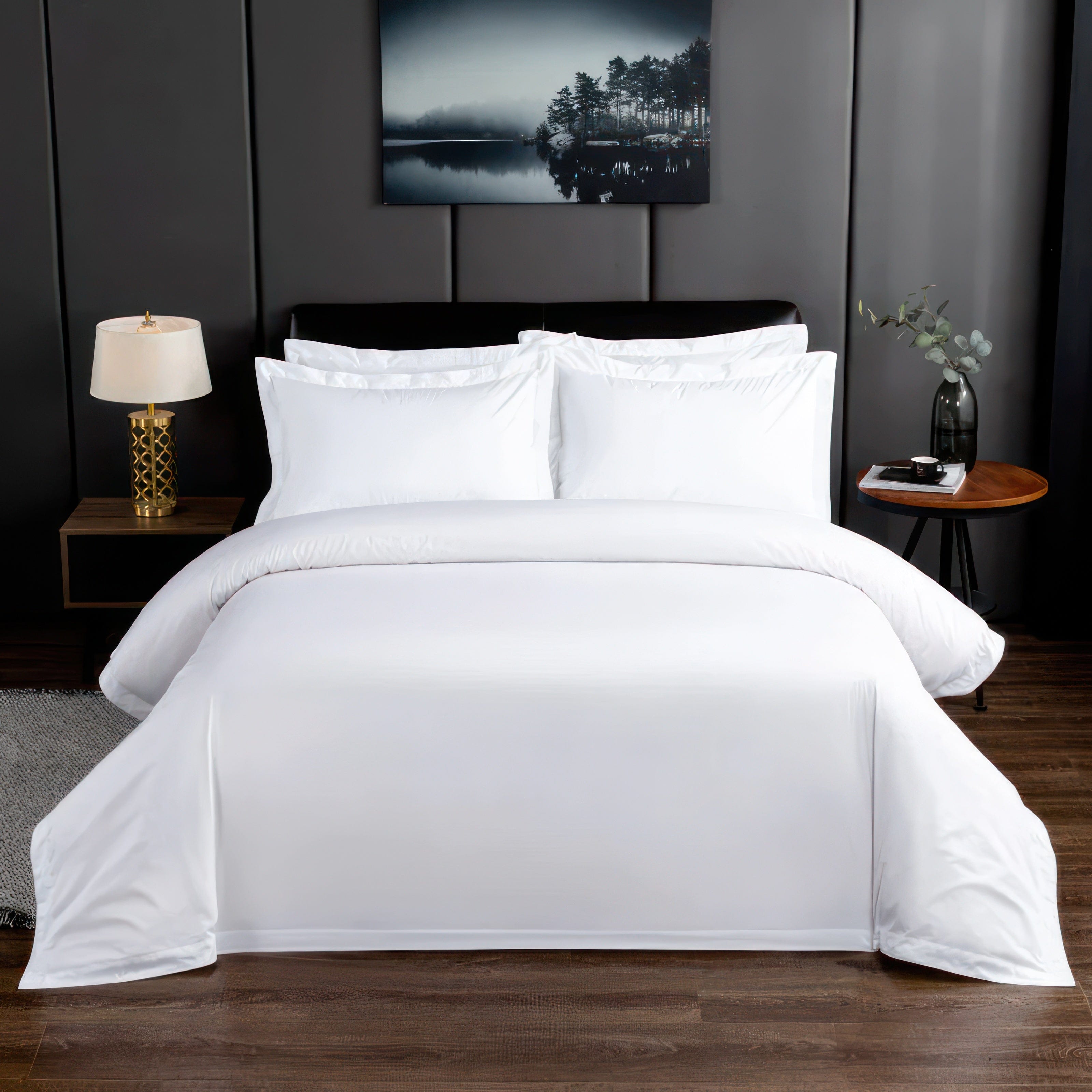 A luxurious white bedding set displayed in a modern bedroom with a sleek black headboard and dark gray walls. The setup includes neatly arranged white pillowcases and a pristine duvet cover, complemented by a round wooden side table with a vase and a cup.

