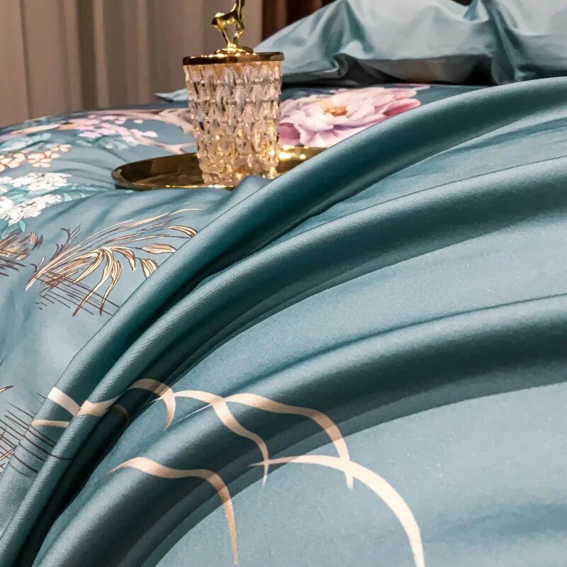 Close-up view of a teal bedding set featuring smooth, luxurious fabric with a slight sheen. The intricate design includes delicate golden lines and floral details, with part of a pink blossom visible. A gold tray with an ornate crystal ornament rests on the duvet, adding a touch of elegance. The fabric's soft folds highlight its silky texture and high quality.

