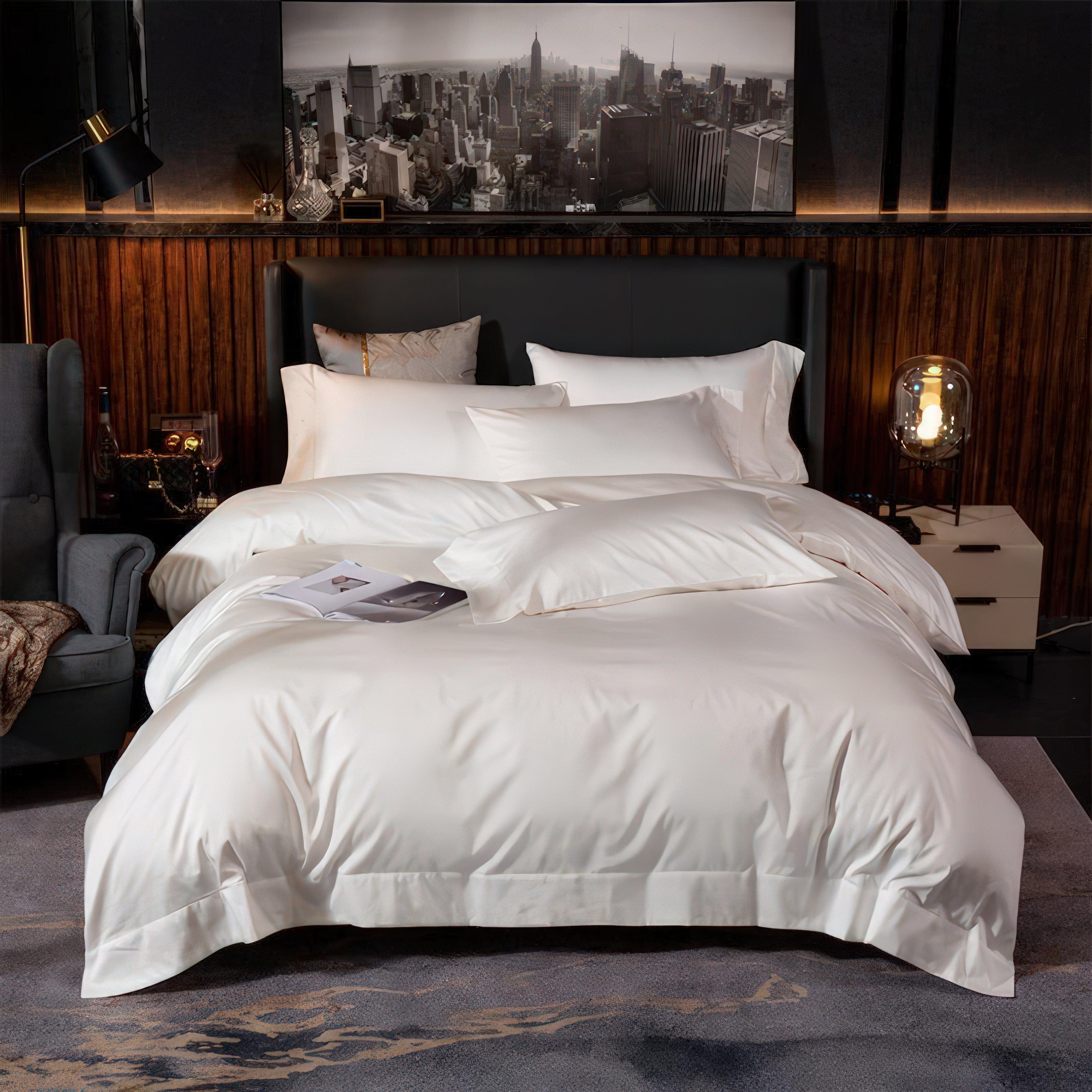 A luxurious white bedding set with a smooth, silky texture is displayed in a sophisticated urban setting. The bed is styled with multiple layers of pillows and a plush duvet, exuding minimalist elegance. The backdrop features a striking black-and-white cityscape photo, warm wooden paneling, and modern bedside decor, including a globe lamp and a velvet armchair, creating a cozy yet refined ambiance.

