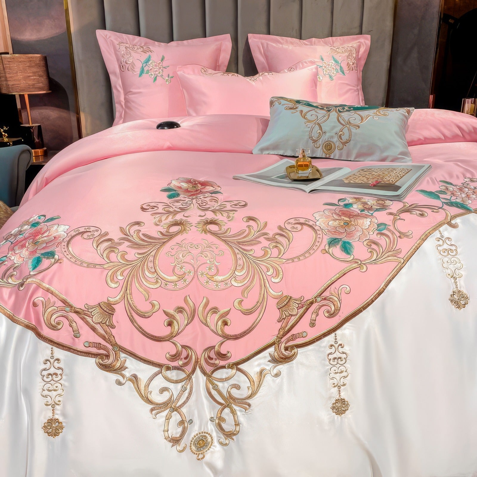 This close-up image highlights the exquisite embroidery of a luxurious bedding set. The pink duvet cover features intricate golden patterns, floral motifs with teal accents, and a white satin border adorned with delicate hanging embellishments. An open book, perfume bottle, and decorative pillow add a touch of sophistication to the setup.

