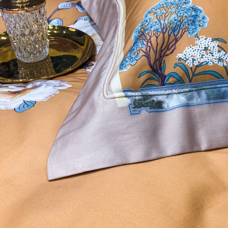 Close-up of an amber pillowcase corner with an embroidered blue tree, white flowers, and a detailed border design. The soft lavender trim frames the pillowcase, set against a smooth amber duvet. A reflective gold tray with a crystal-textured glass rests beside it.

