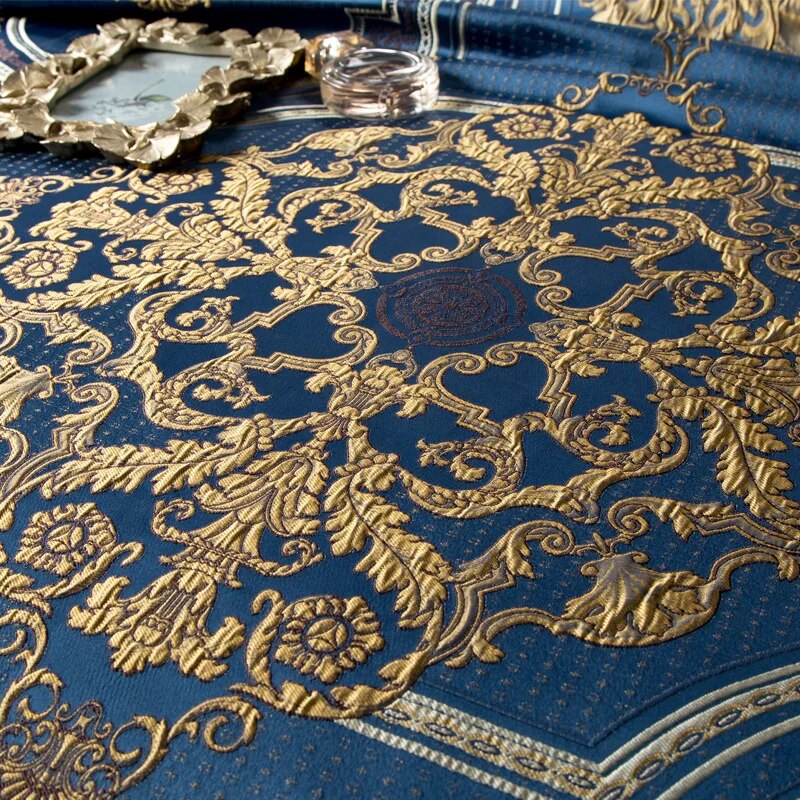 Close-up of an ornate blue fabric featuring intricate gold embroidery in an elaborate baroque-style pattern. The luxurious design includes swirling vines, floral motifs, and symmetrical details, with fine gold stitching creating a regal, opulent appearance. A decorative mirror frame and a glass perfume bottle are subtly placed in the background, enhancing the elegance of the scene.

