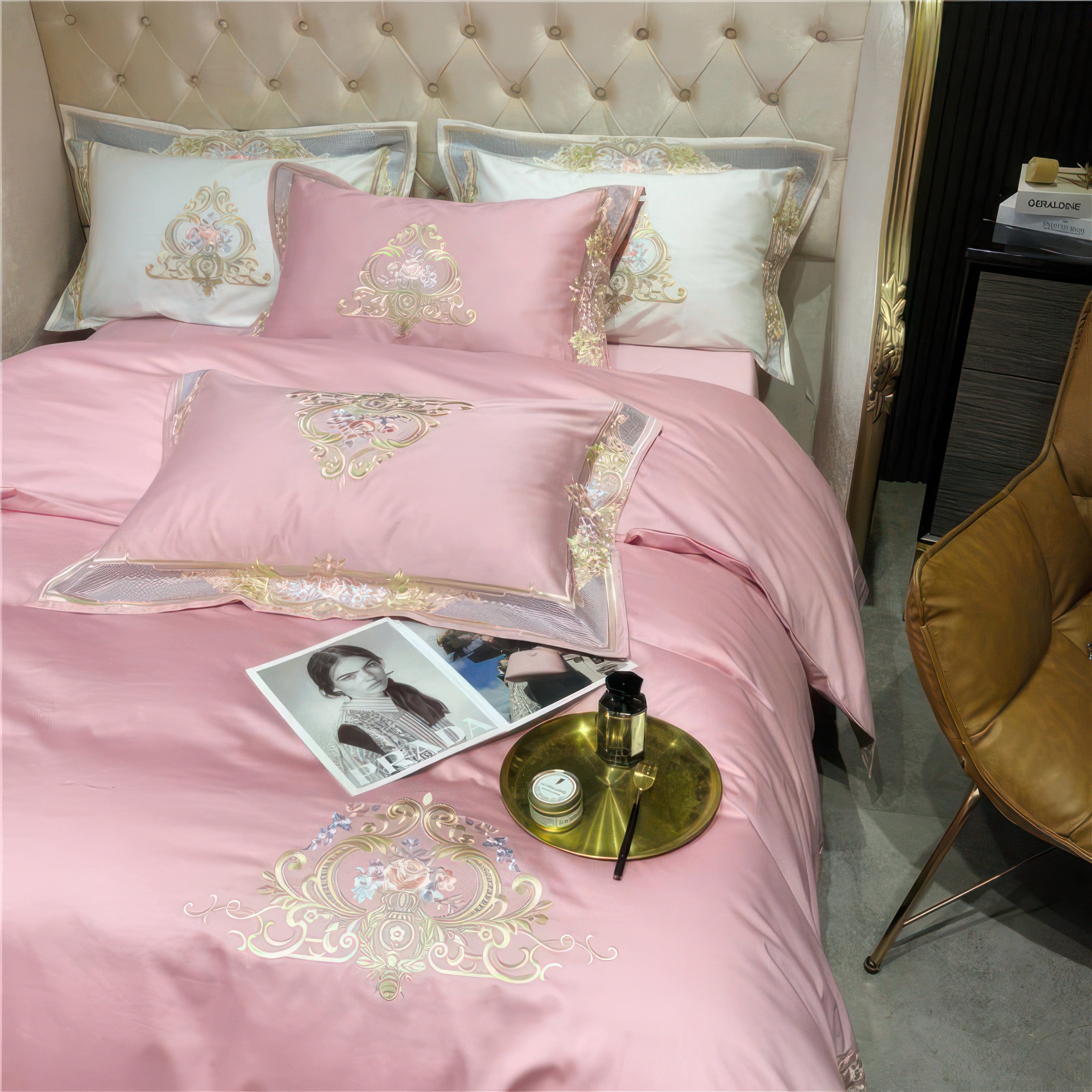 A close-up view of a pink bedding set with intricate gold embroidery and floral motifs, complemented by a decorative golden tray holding a candle, perfume, and a black reed diffuser. The bedding features layers of elegance with embroidered white pillows in the background, while the scene is accented with a luxury tufted headboard and sophisticated side decor, emphasizing the opulence of the setup.

