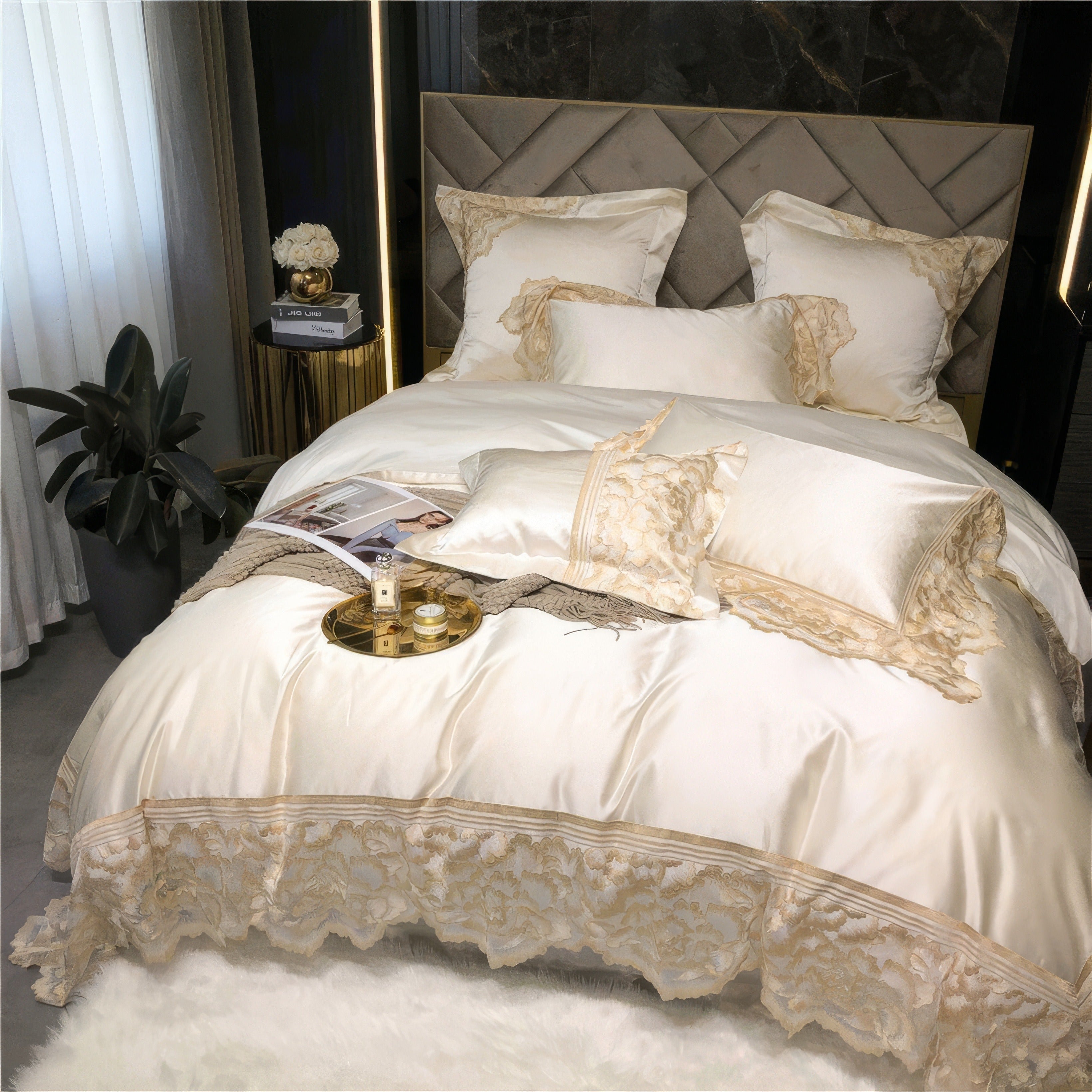 White satin bedding set with gold lace detailing, featuring decorative pillows, a soft throw, and a golden tray with perfume bottles, styled against a tufted headboard and marble wall.

