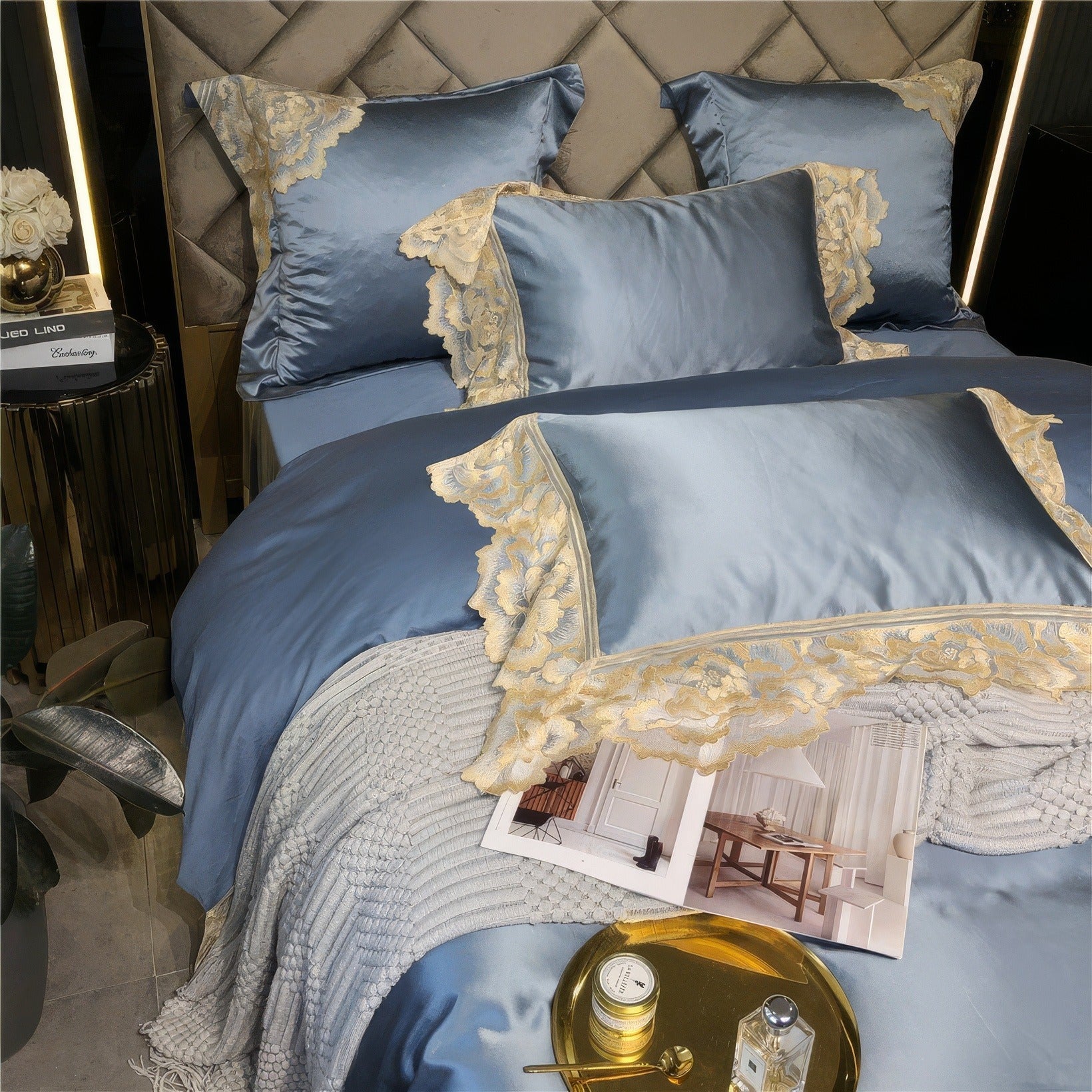 Blue satin bedding with gold lace details, decorative pillows, a textured white knit blanket, a gold tray holding a candle and perfume, and an open interior design magazine.

