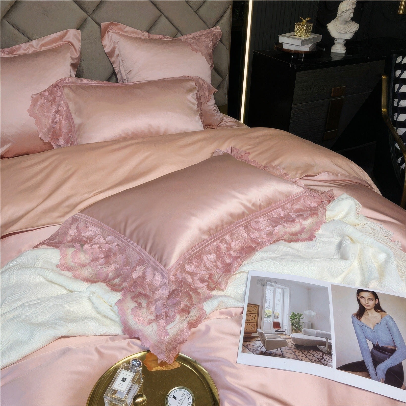 Close-up of a pink satin bedding set with intricate lace trim on a pillow, a white throw blanket, and an open magazine featuring interior design and a model in a blue top, with a gold tray holding perfume and a candle.

