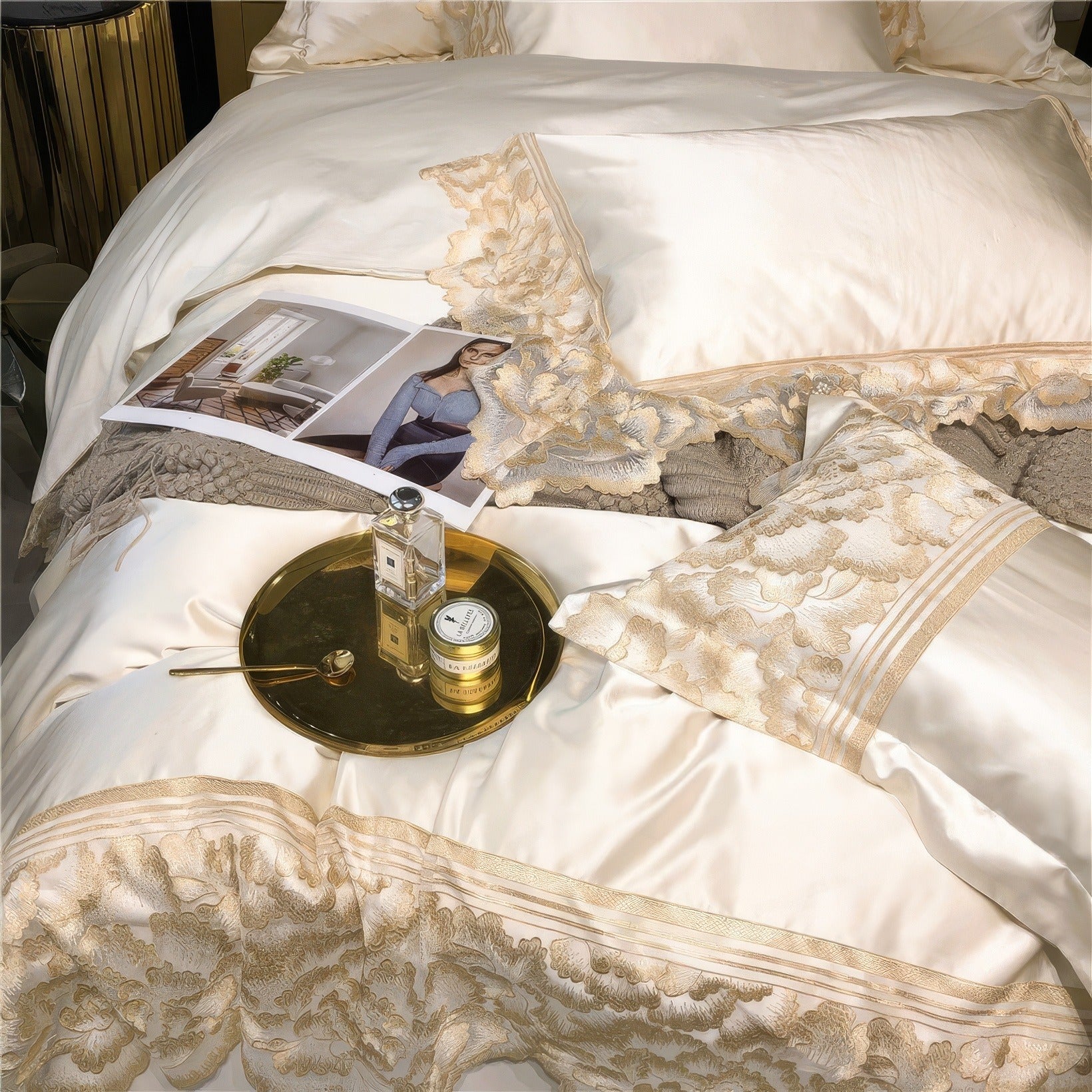 Close-up of white satin bedding with intricate gold lace detailing, a decorative pillow, a magazine, and a gold tray holding perfume and a small jar, styled on a soft, textured blanket.

