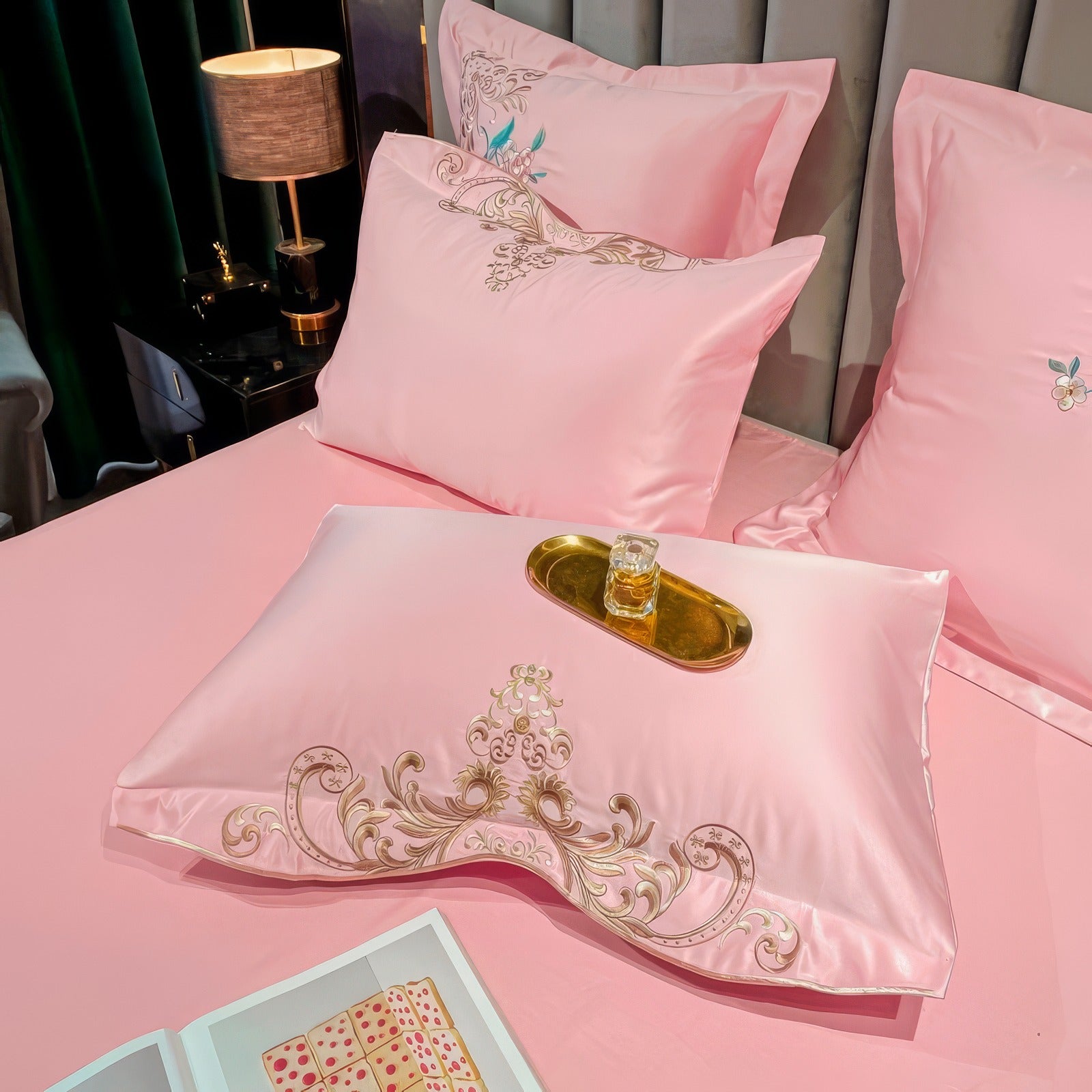 Close-up of a pink bedding set with intricately embroidered pillowcases featuring ornate golden scrollwork and floral accents in pink and teal. The centerpiece includes a decorative light blue pillow with matching embroidery, and a golden tray holding a small glass perfume bottle placed on the duvet. An open book rests nearby, adding a cozy and luxurious touch to the scene.

