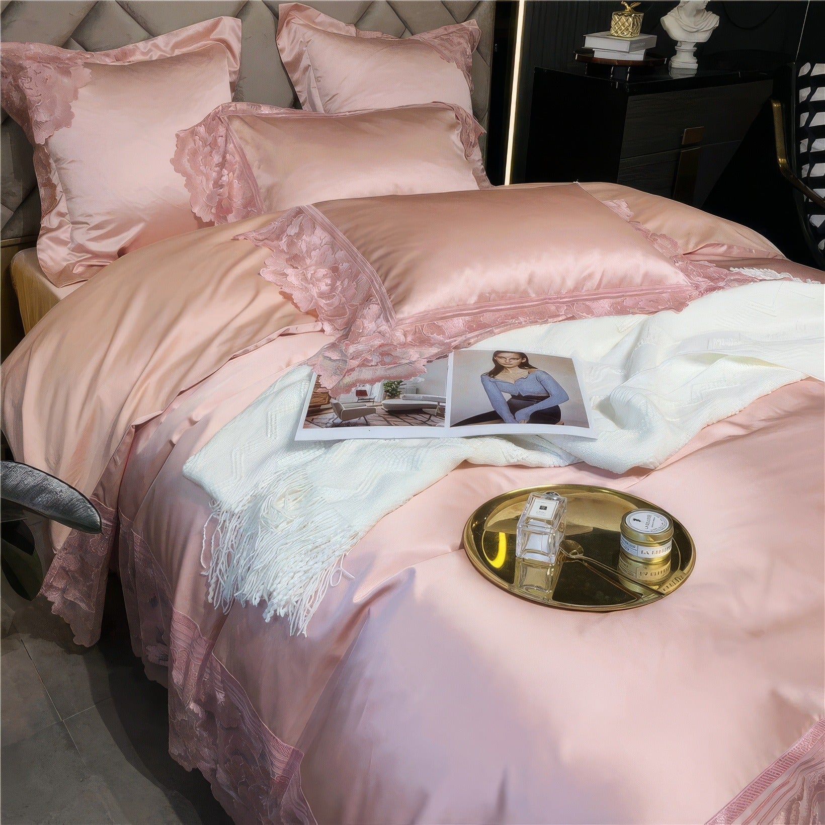 Close-up of a pink satin bedding set with delicate lace details on pillows and edges, featuring a white throw blanket, an open magazine, and a gold tray holding a candle and perfume bottle.

