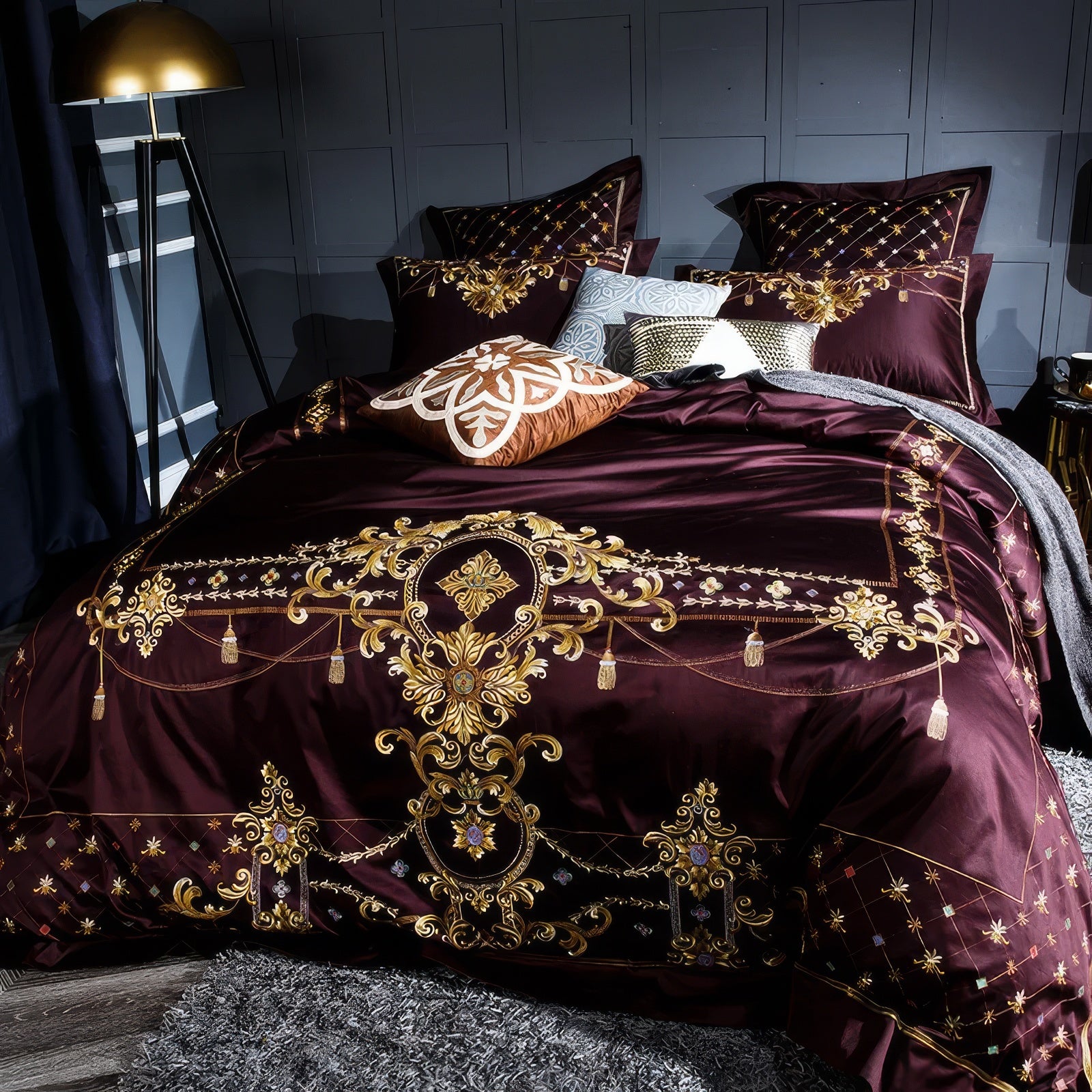 Maroon bedding set adorned with intricate gold embroidery, baroque-inspired patterns, and tassel details, paired with accent pillows featuring unique geometric and textured designs. Set in a modern dark-themed room with elegant gold lighting and a luxurious rug.

