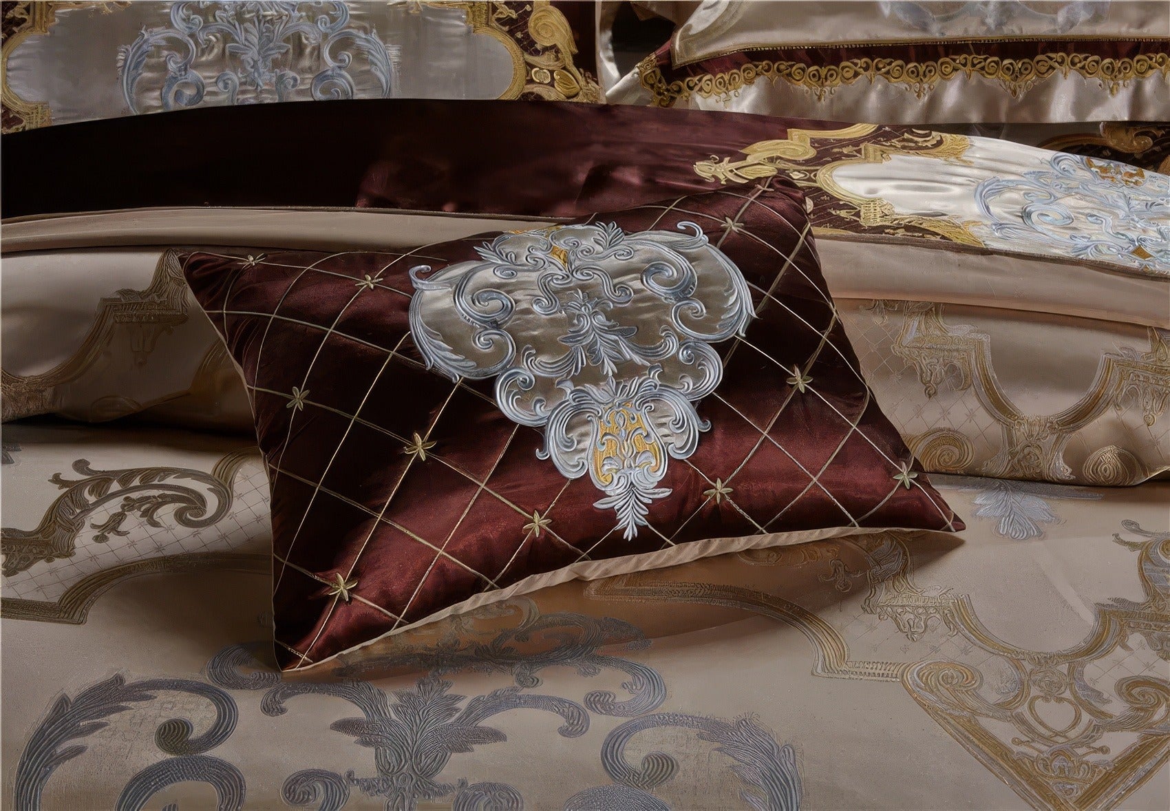 A close-up view of a decorative pillow in luxurious burgundy fabric adorned with an elegant silver and gold embroidery pattern. The pillow features a quilted design with delicate golden star accents at each intersection, adding texture and sophistication. The bedding beneath complements the design with intricate patterns in champagne, silver, and gold tones, creating an opulent and harmonious aesthetic.

