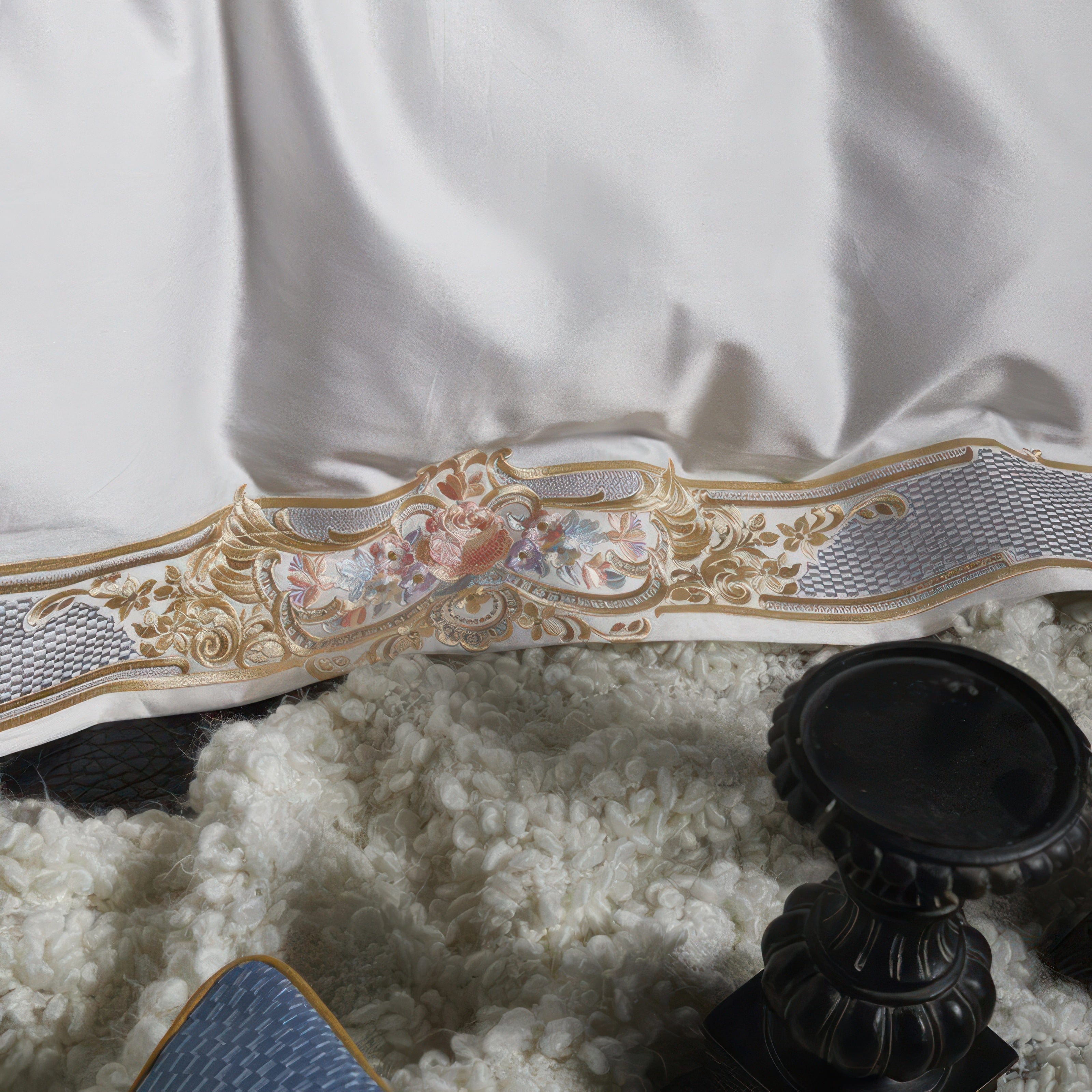 A close-up of a luxurious white bedding adorned with detailed gold and pastel embroidery. The intricate design includes floral accents and decorative borders, complemented by a silver latticework pattern. The bedding rests on a textured white blanket, with a black decorative item partially visible, adding a touch of elegance to the setting.

